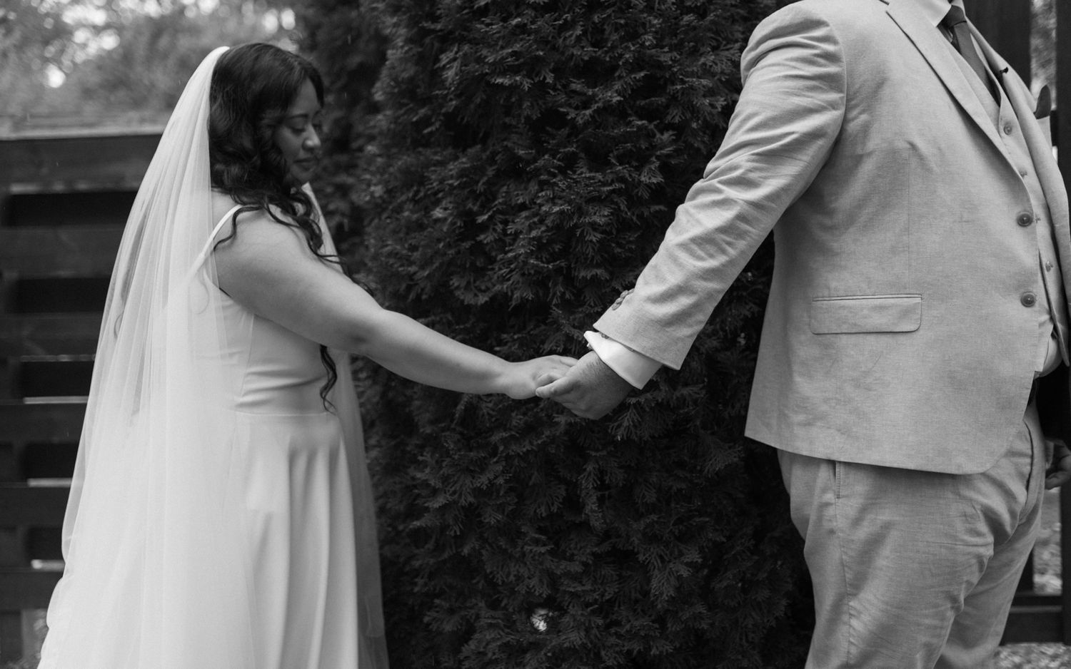 A couple in wedding attire holding hands in black and white with a hedge backdrop.