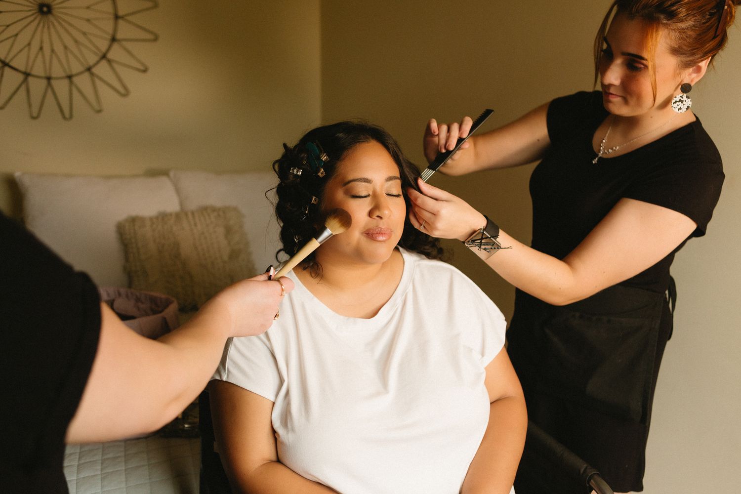A hairstylist wearing black styles and arranges their client's dark curly hair in a sunlit room.