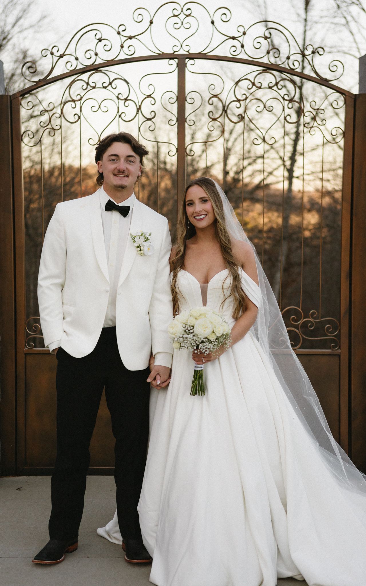 A romantic wedding photo of a couple in front of an ornate iron gate at sunset.