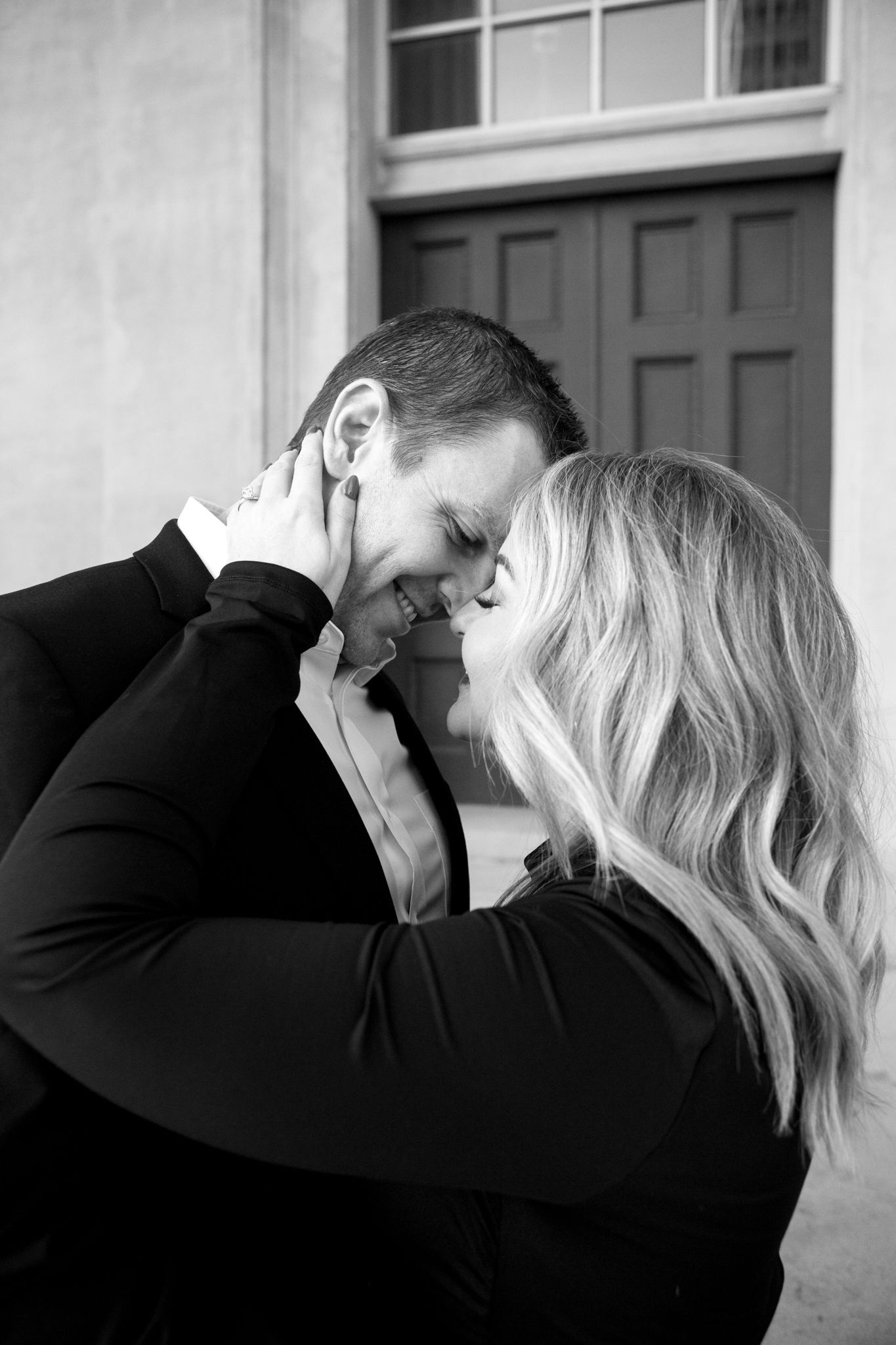 A tender black and white portrait captures an intimate moment between a couple outside a building.