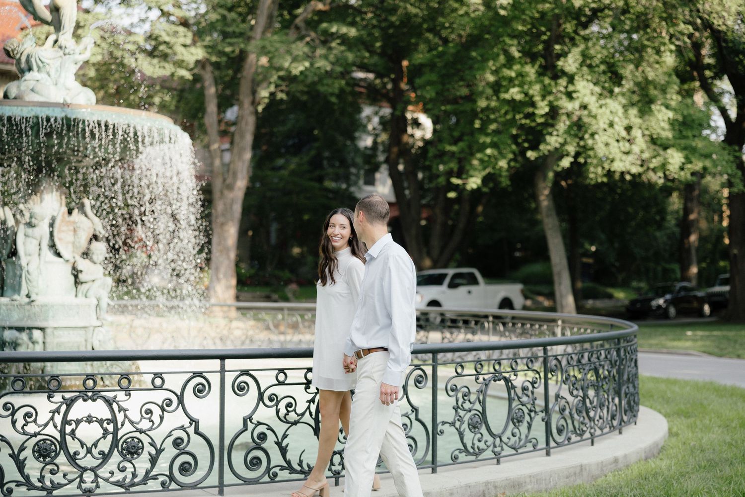 A couple in white outfits walks near an ornate fountain in a lush green park on a sunny day.