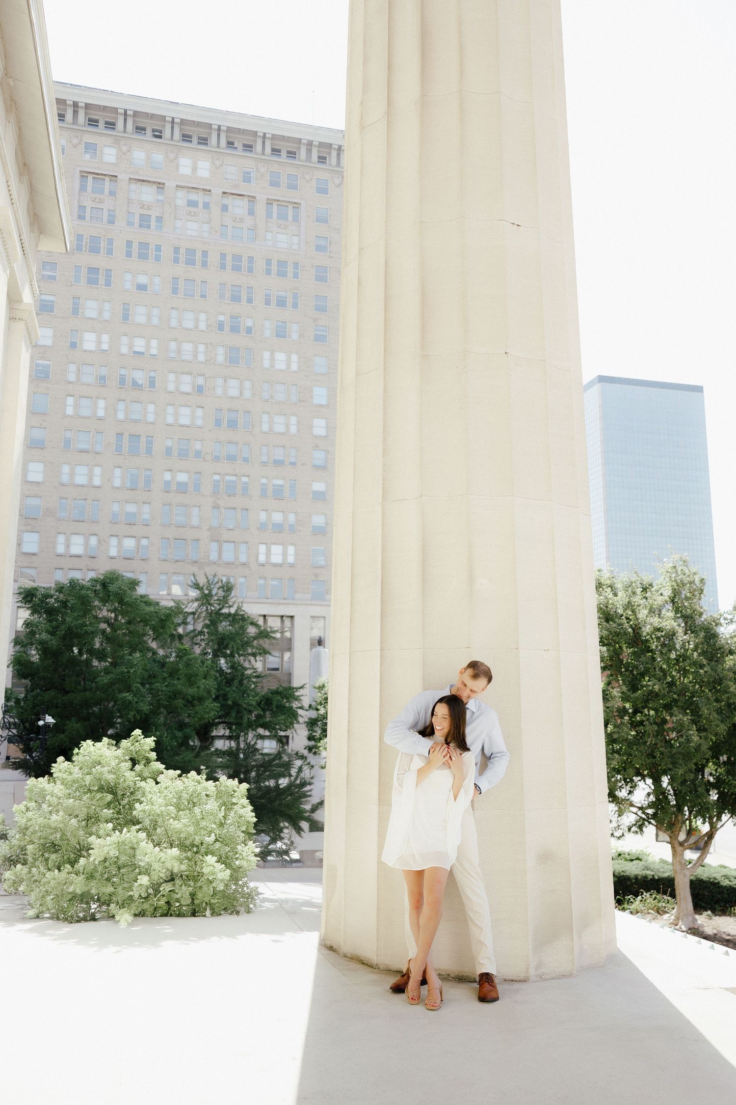 A couple poses romantically between large white columns with city buildings visible in the background.