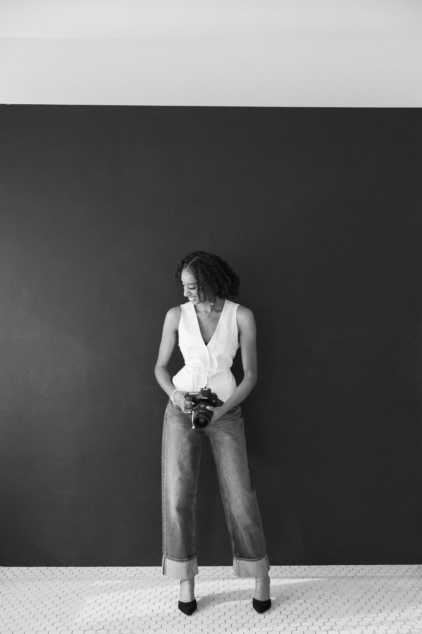 Black and white photo of a stylish woman in wide-leg jeans holding a camera against a dark wall.