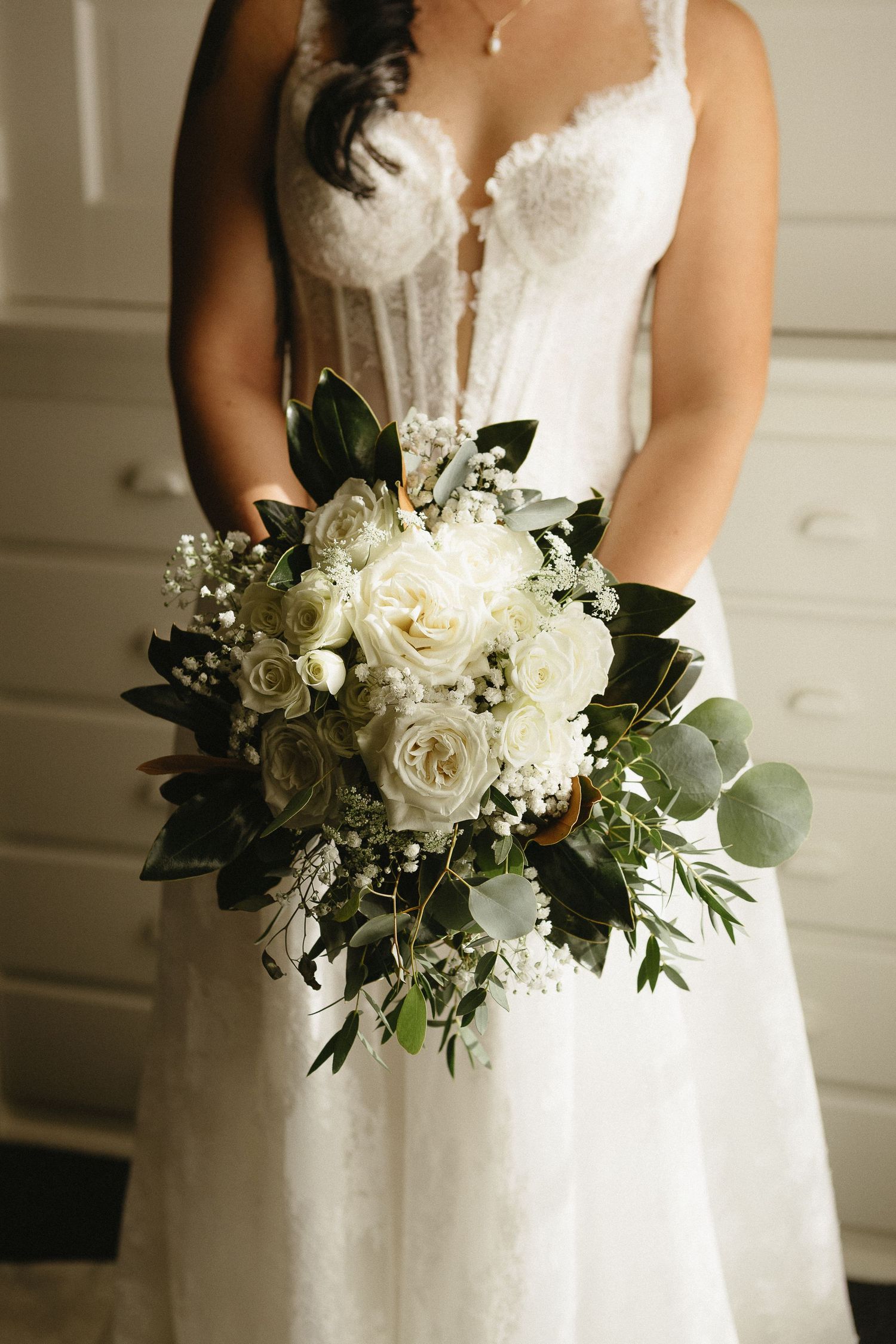 White bridal bouquet with roses and eucalyptus leaves held against an elegant wedding gown.