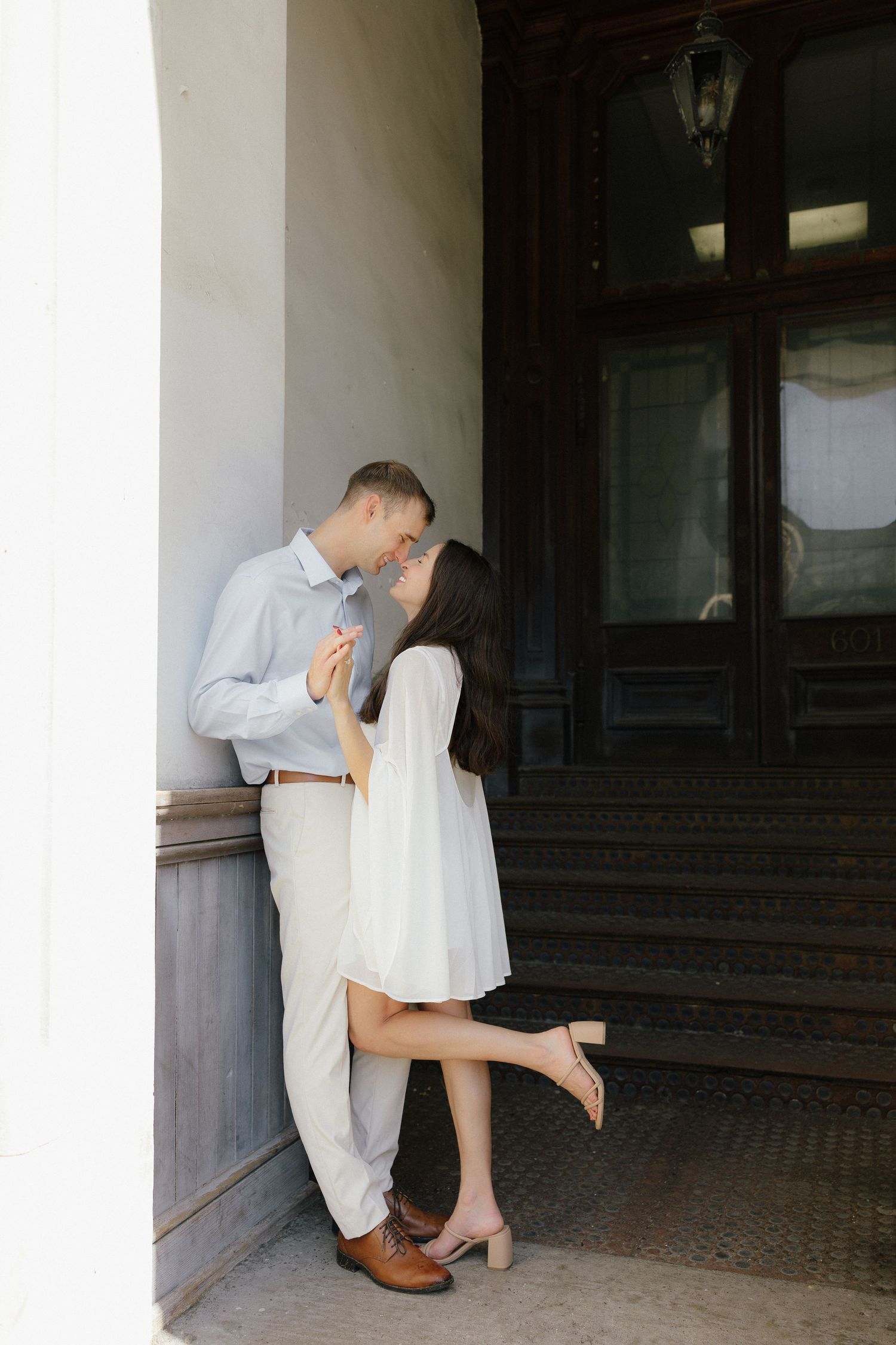 Couple sharing a kiss near a vintage doorway, woman in white dress with one heel lifted romantically.