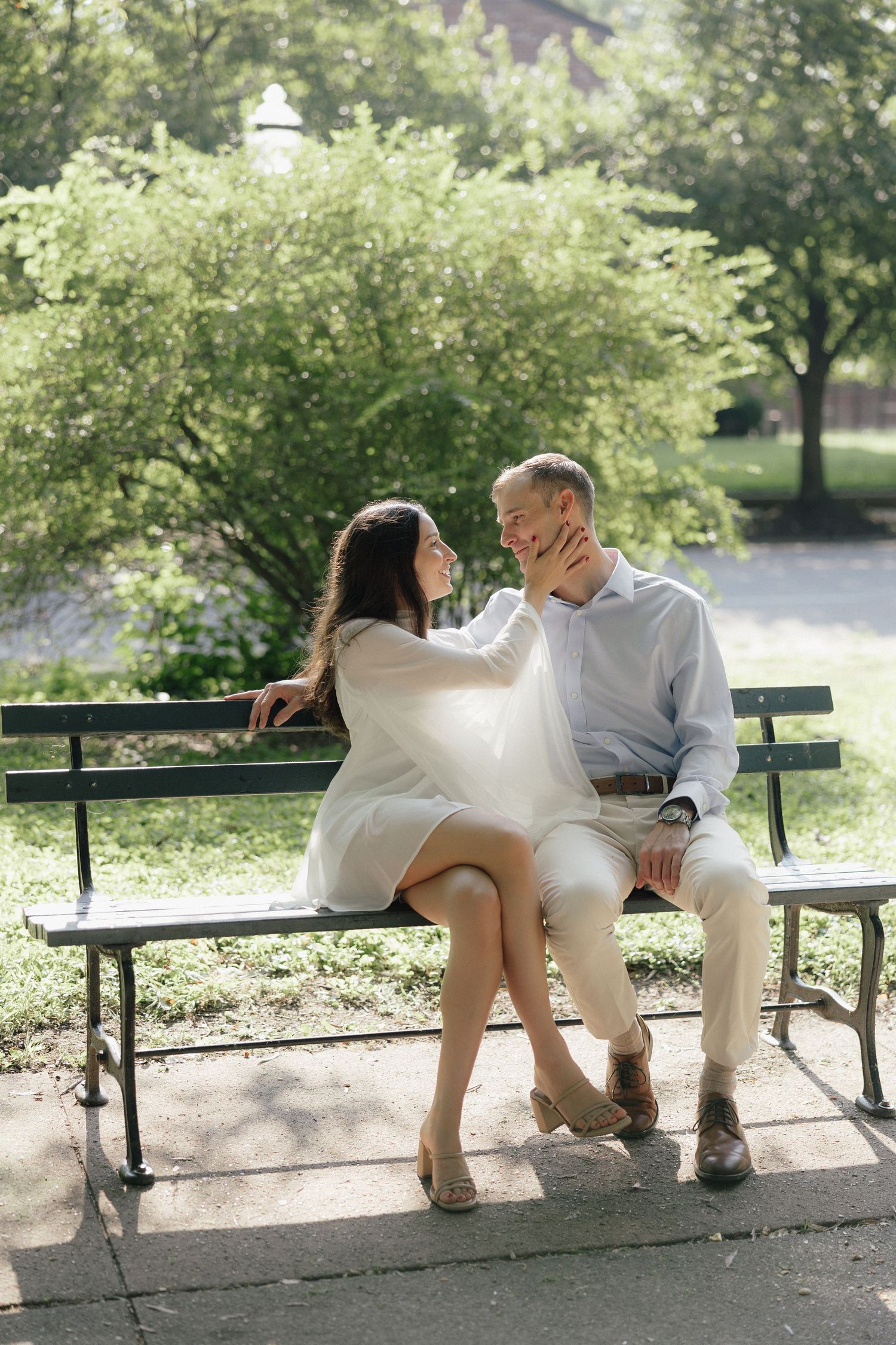 Couple sitting on a park bench, woman in white dress touching mans face, surrounded by lush green trees.