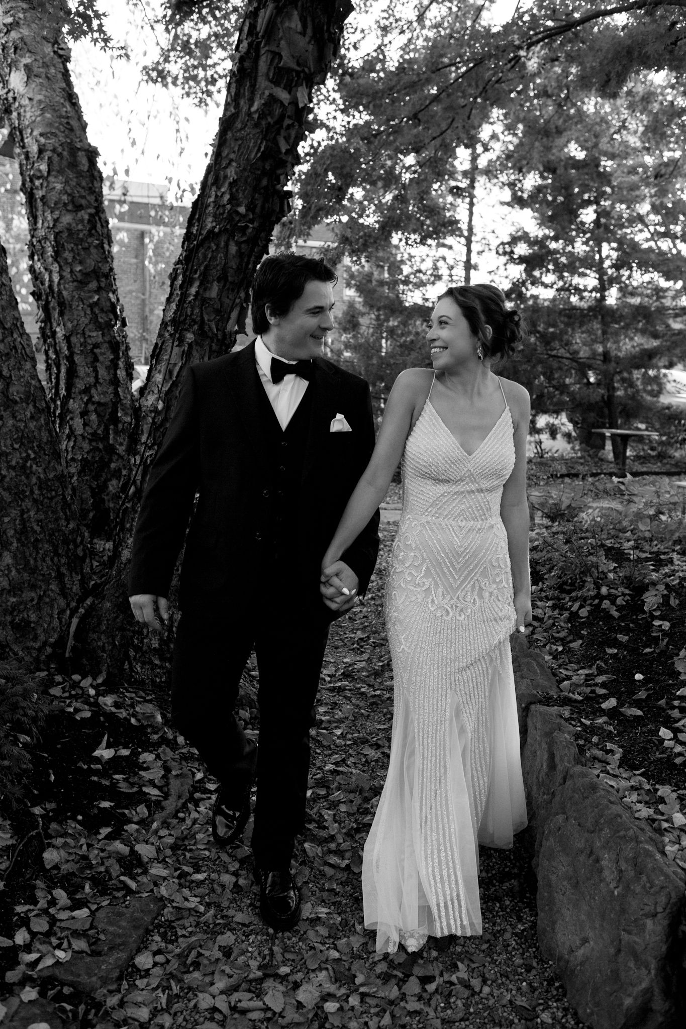 Black and white portrait of a couple in wedding attire walking together through an autumn garden.