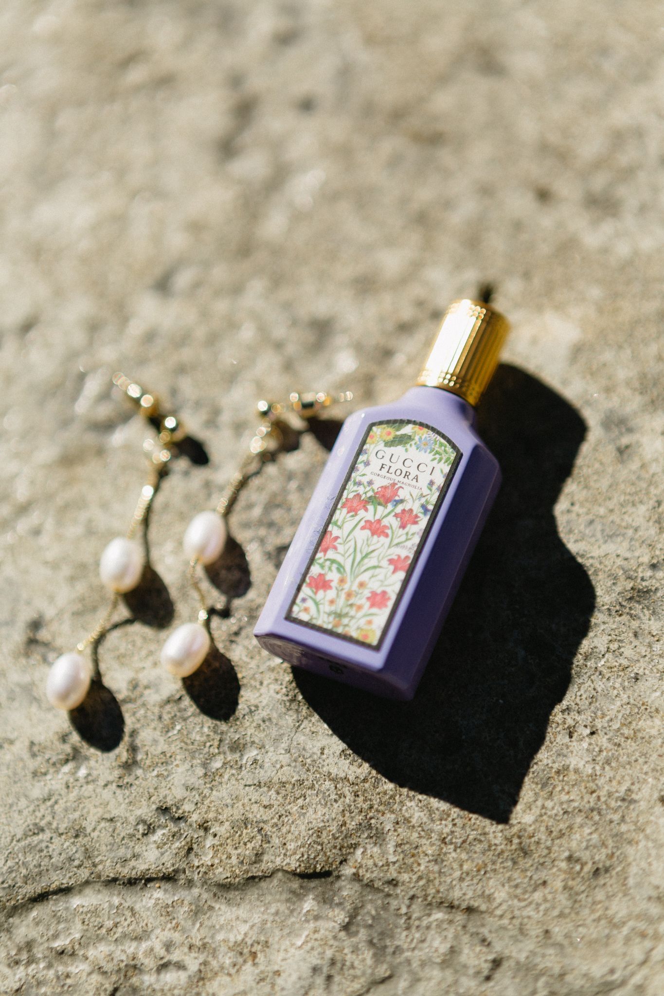 Pearl earrings and purple perfume bottle arranged artistically on textured stone surface.