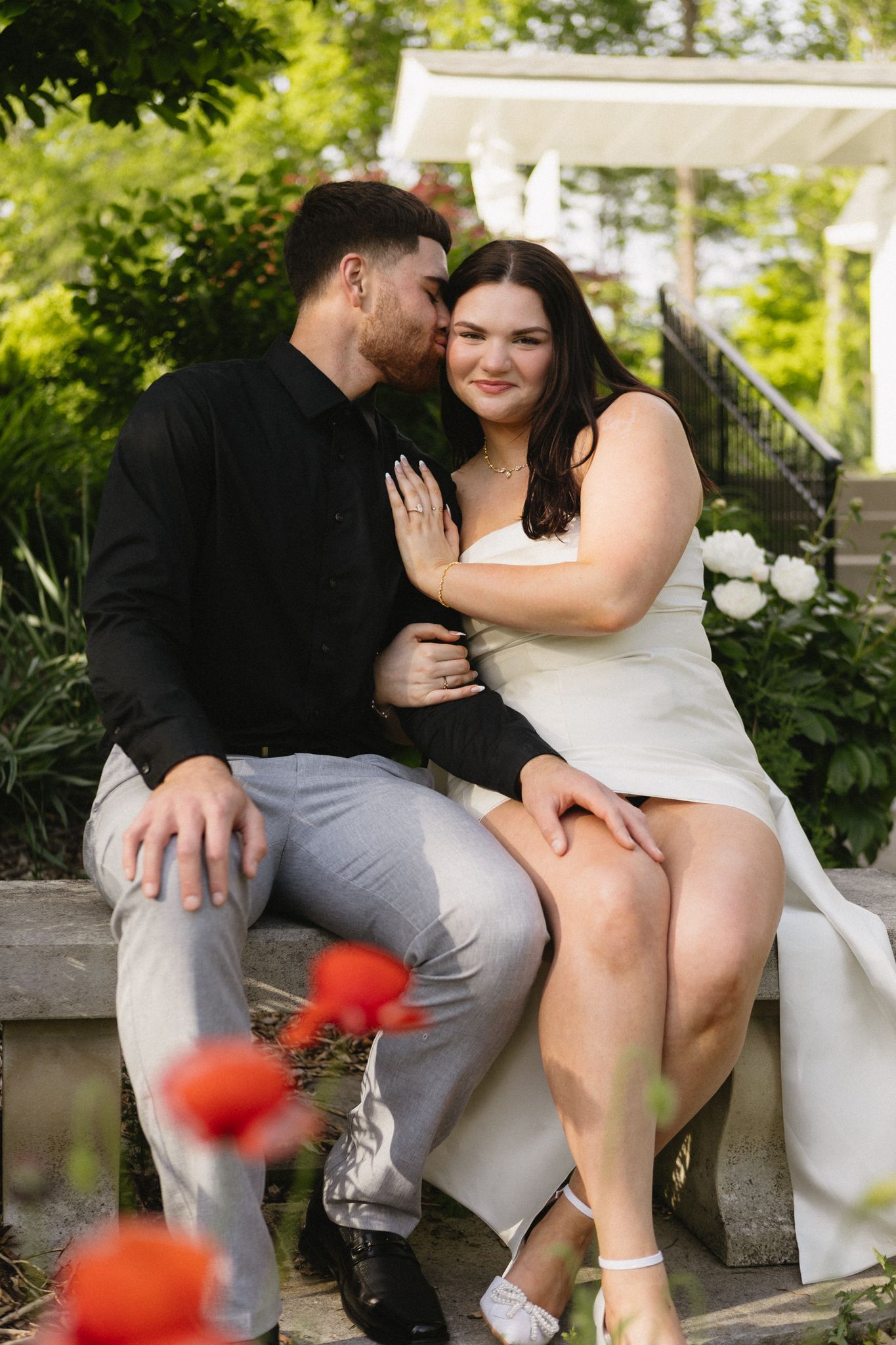 A romantic photoshoot captures intimate moments between a couple on a garden bench with red flowers in the foreground.