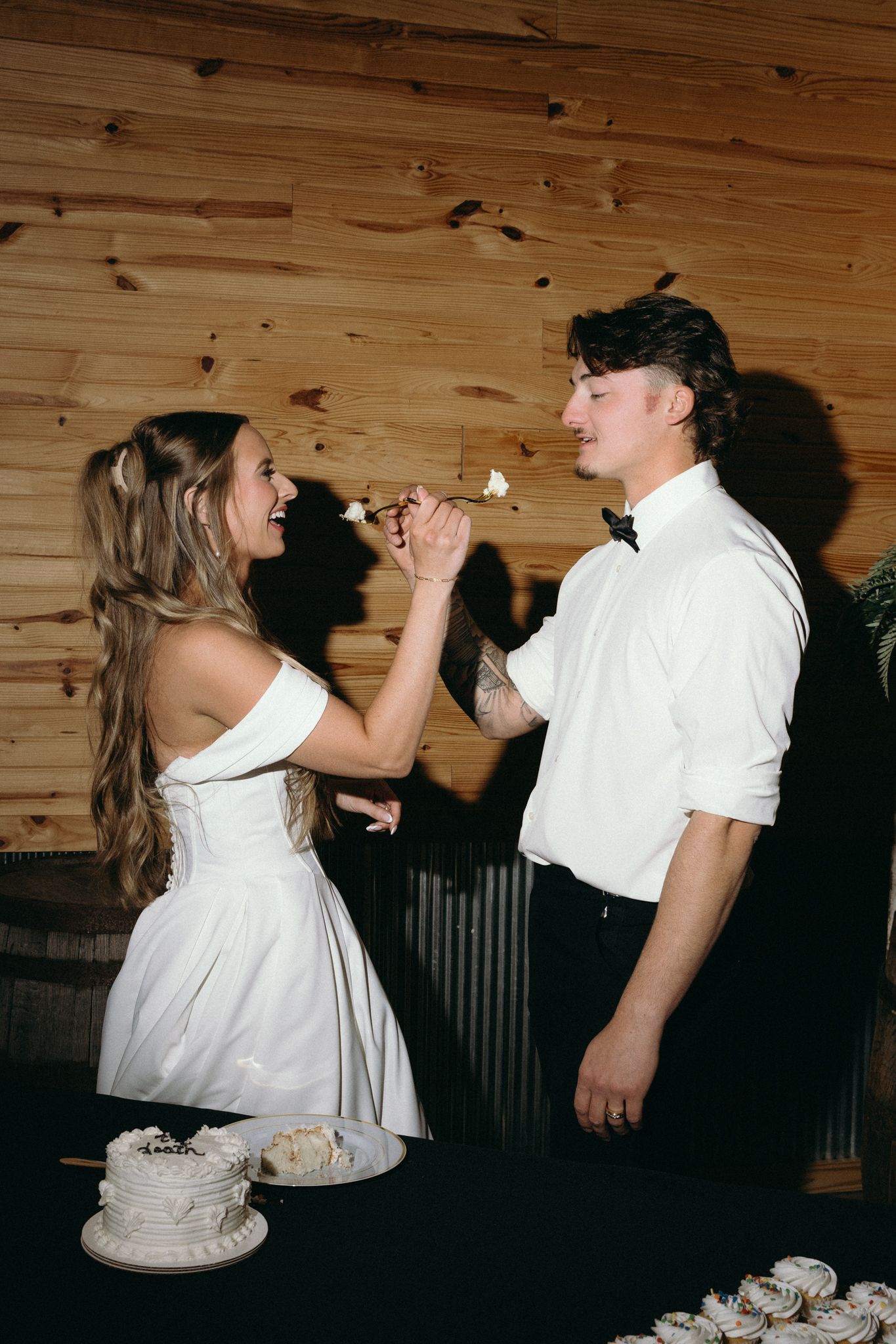Newlyweds share a playful moment feeding each other wedding cake in a rustic wooden venue.
