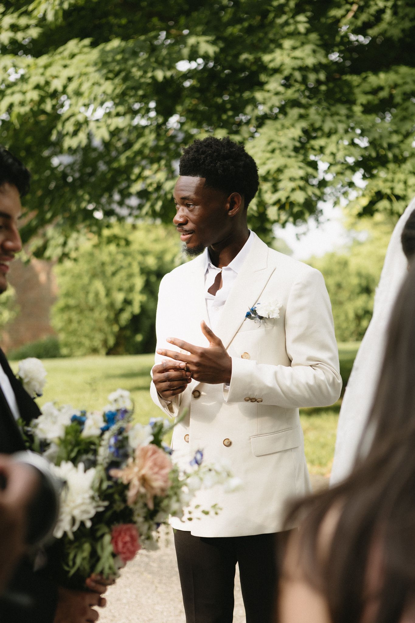 A groom in a crisp white double-breasted jacket speaks during an outdoor wedding ceremony.
