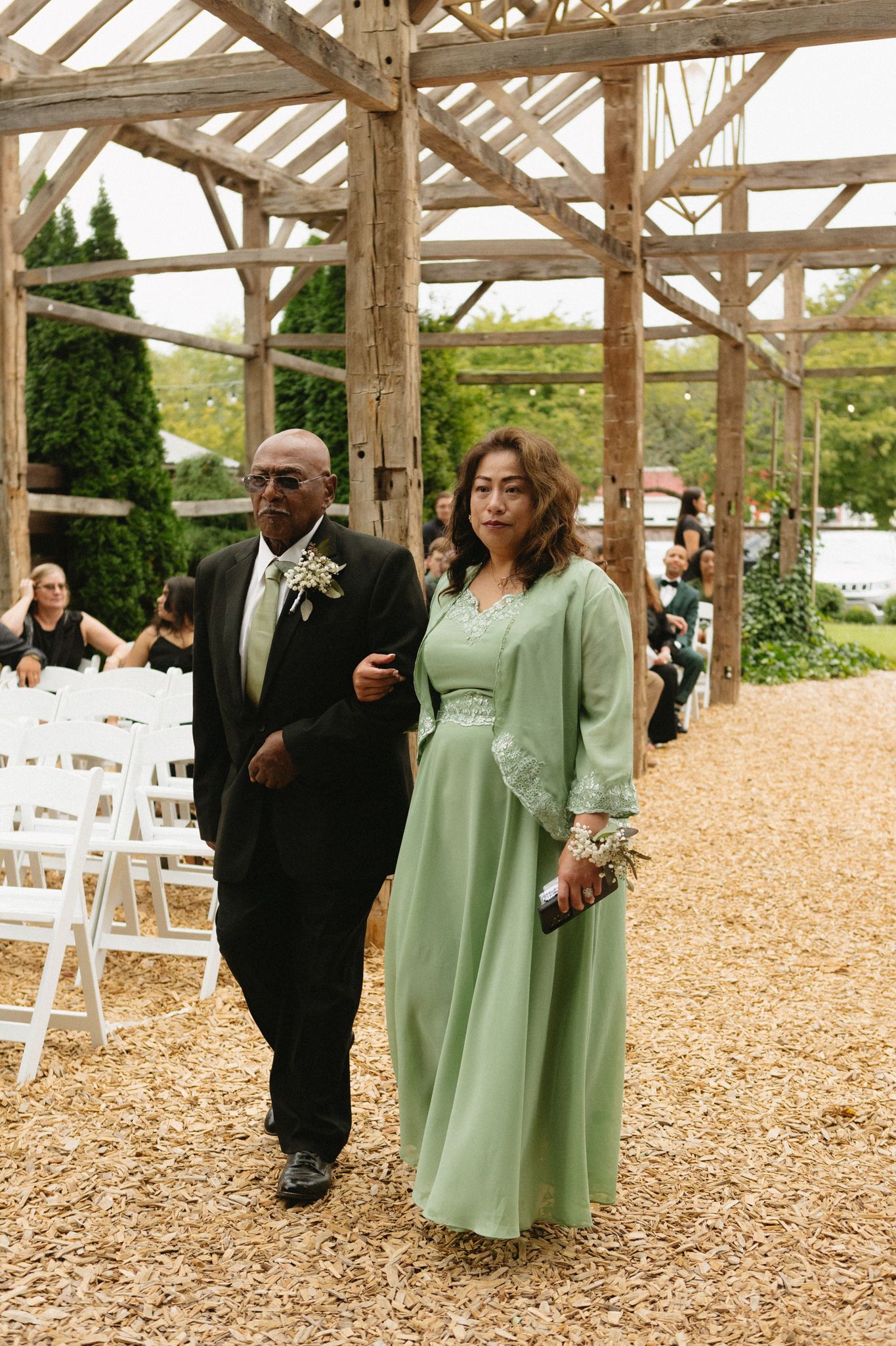 Wedding attendees walk down an aisle under a rustic wooden pergola with white chairs and greenery in the background.