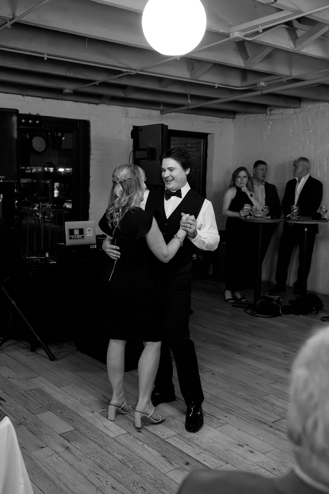 Couples dancing at an indoor wedding reception with hardwood floors and industrial lighting.