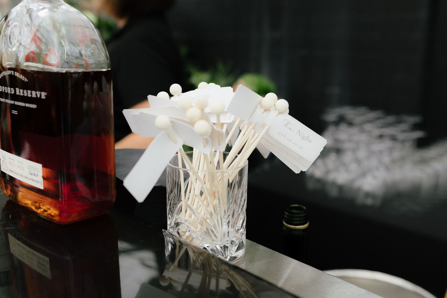 Crystal vase with white cocktail stirrers and bourbon bottle on black reflective bar surface.