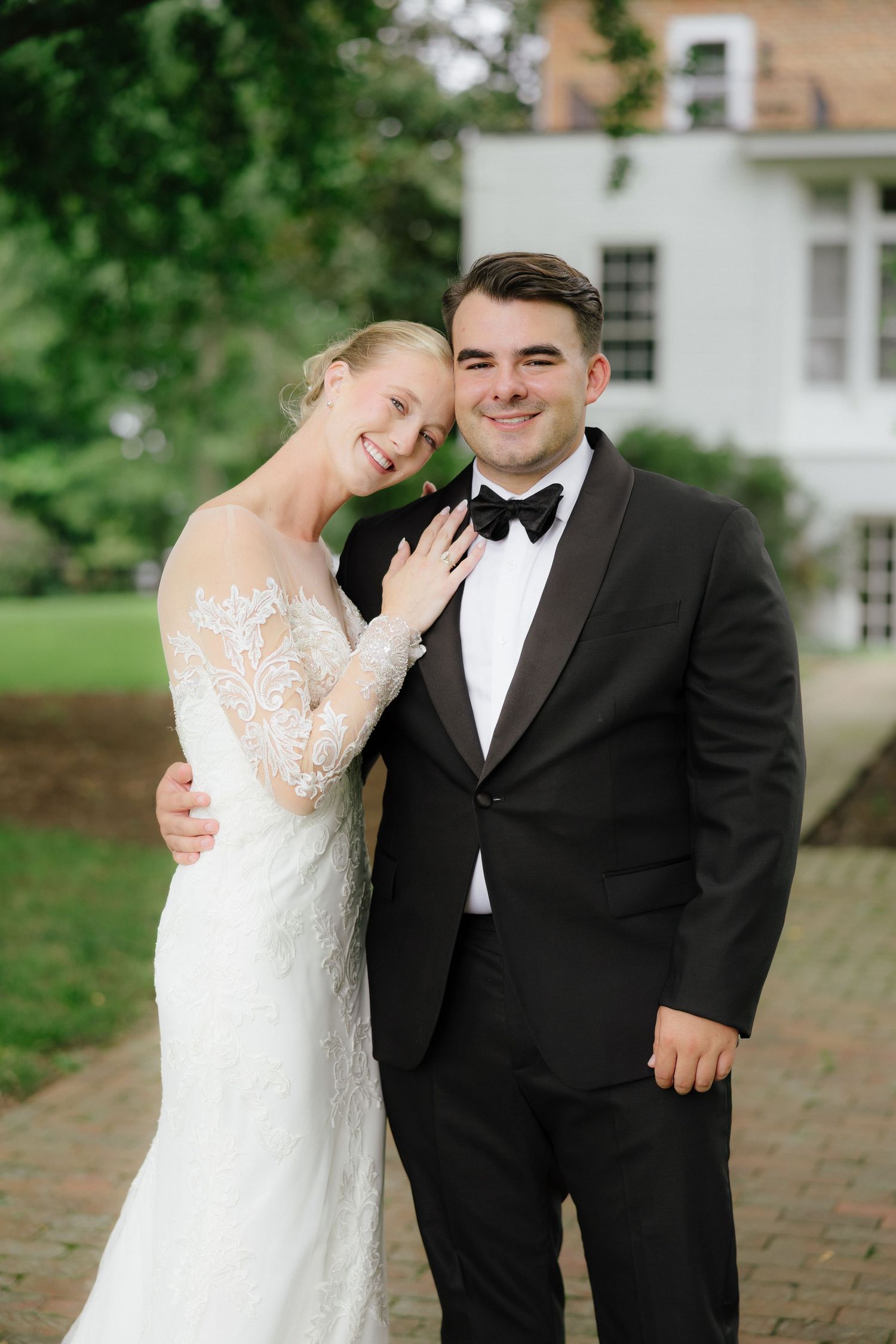 Bride and groom pose together outdoors, she wears lace gown, he wears black tuxedo.