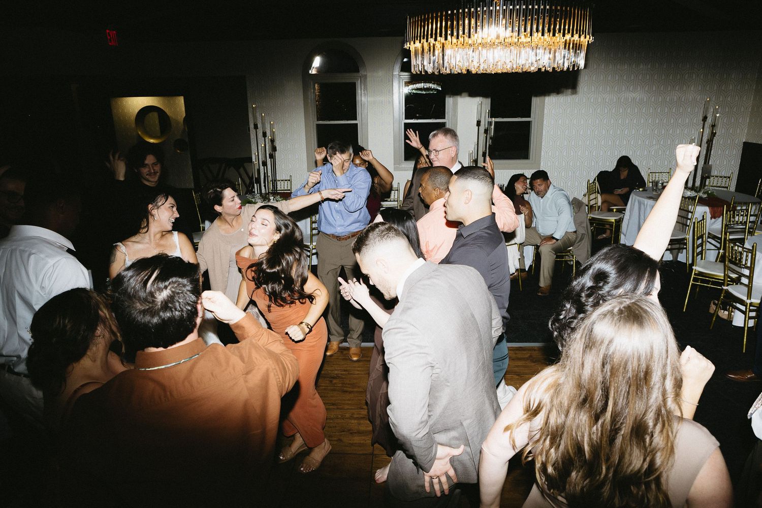 Guests dancing and celebrating at a dimly lit wedding reception under ornate lighting.