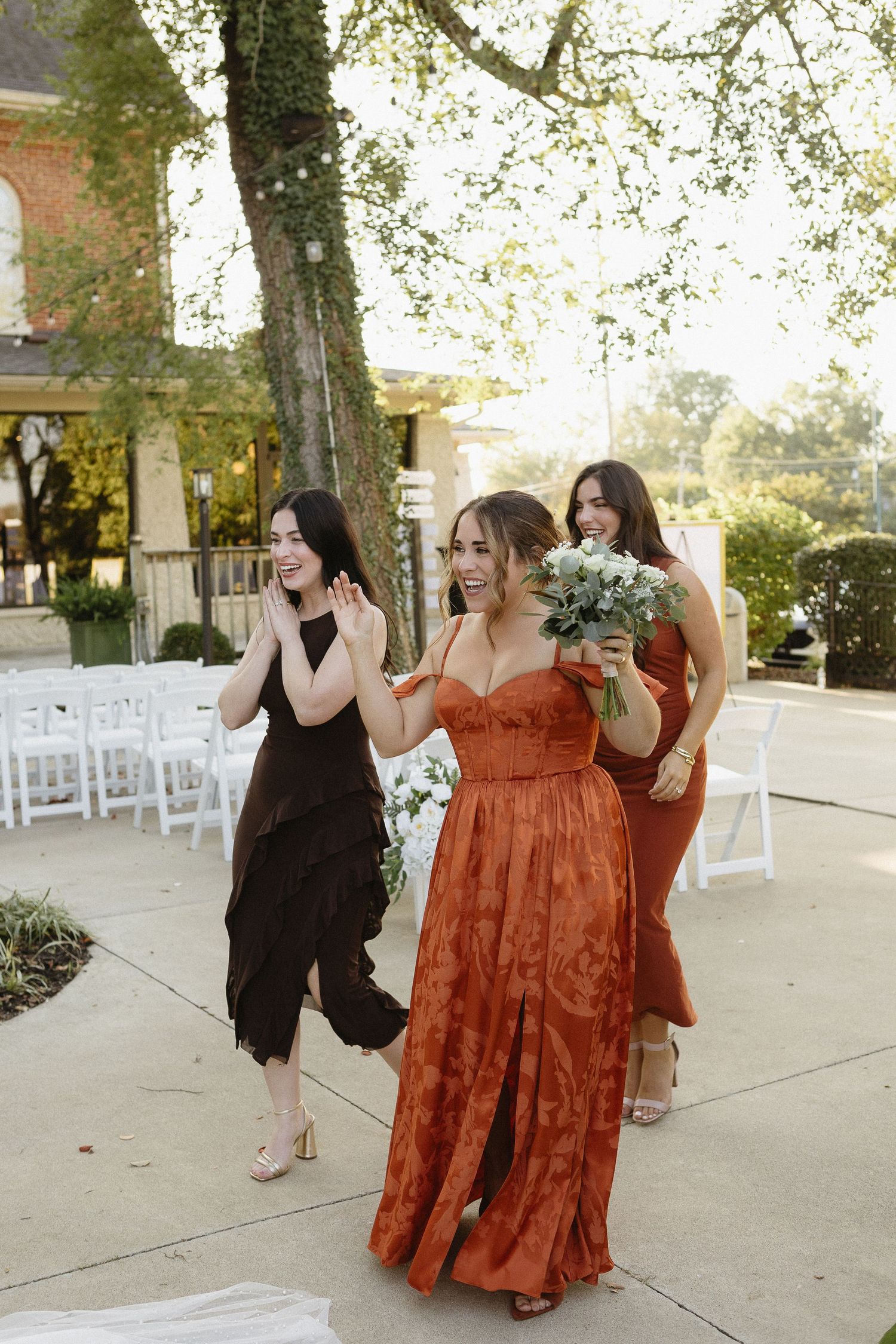 Bridesmaids in rust-colored and black dresses share candid moments at an outdoor wedding venue.