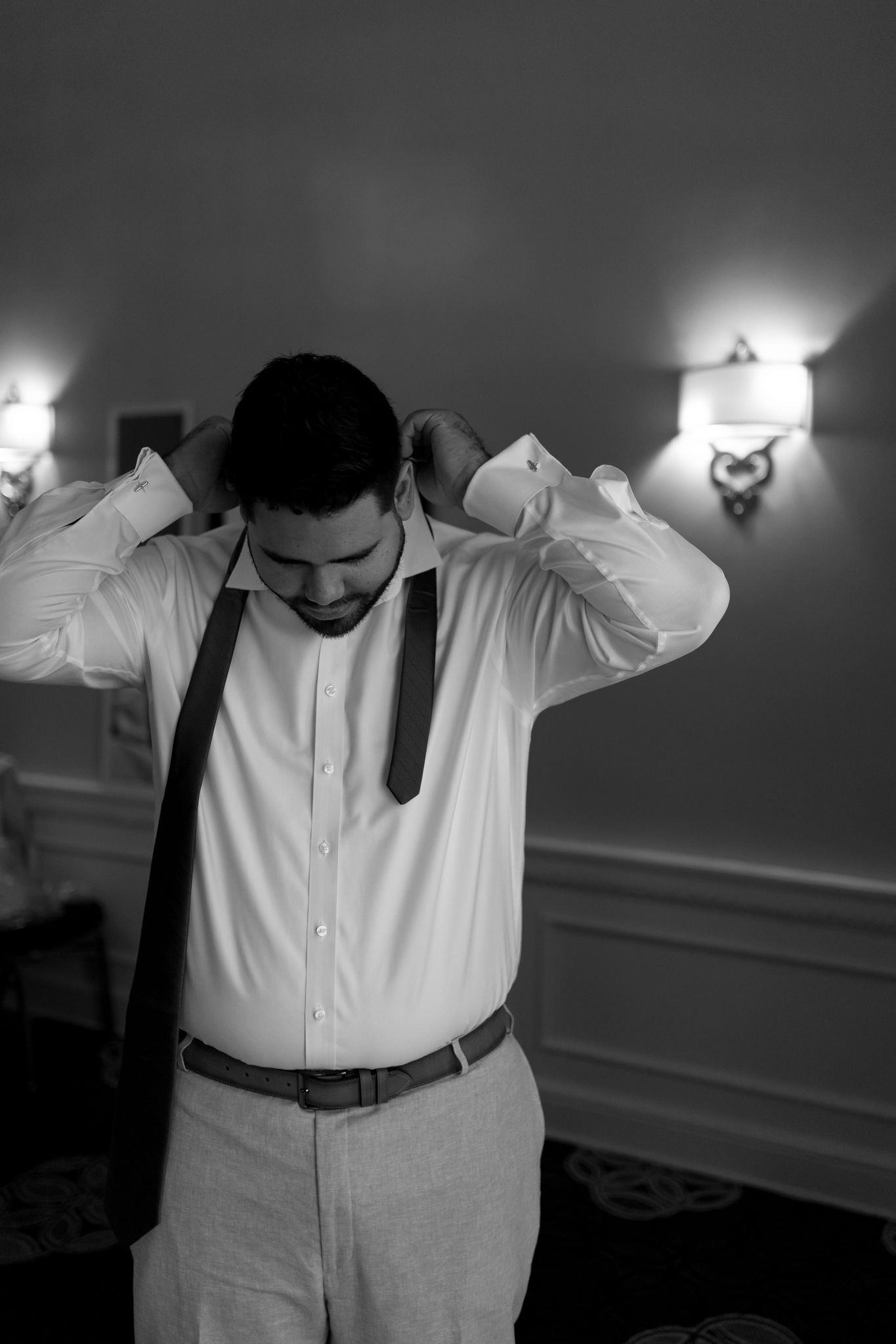 A person in formal attire adjusts their white dress shirt while wearing suspenders in a dimly lit room.