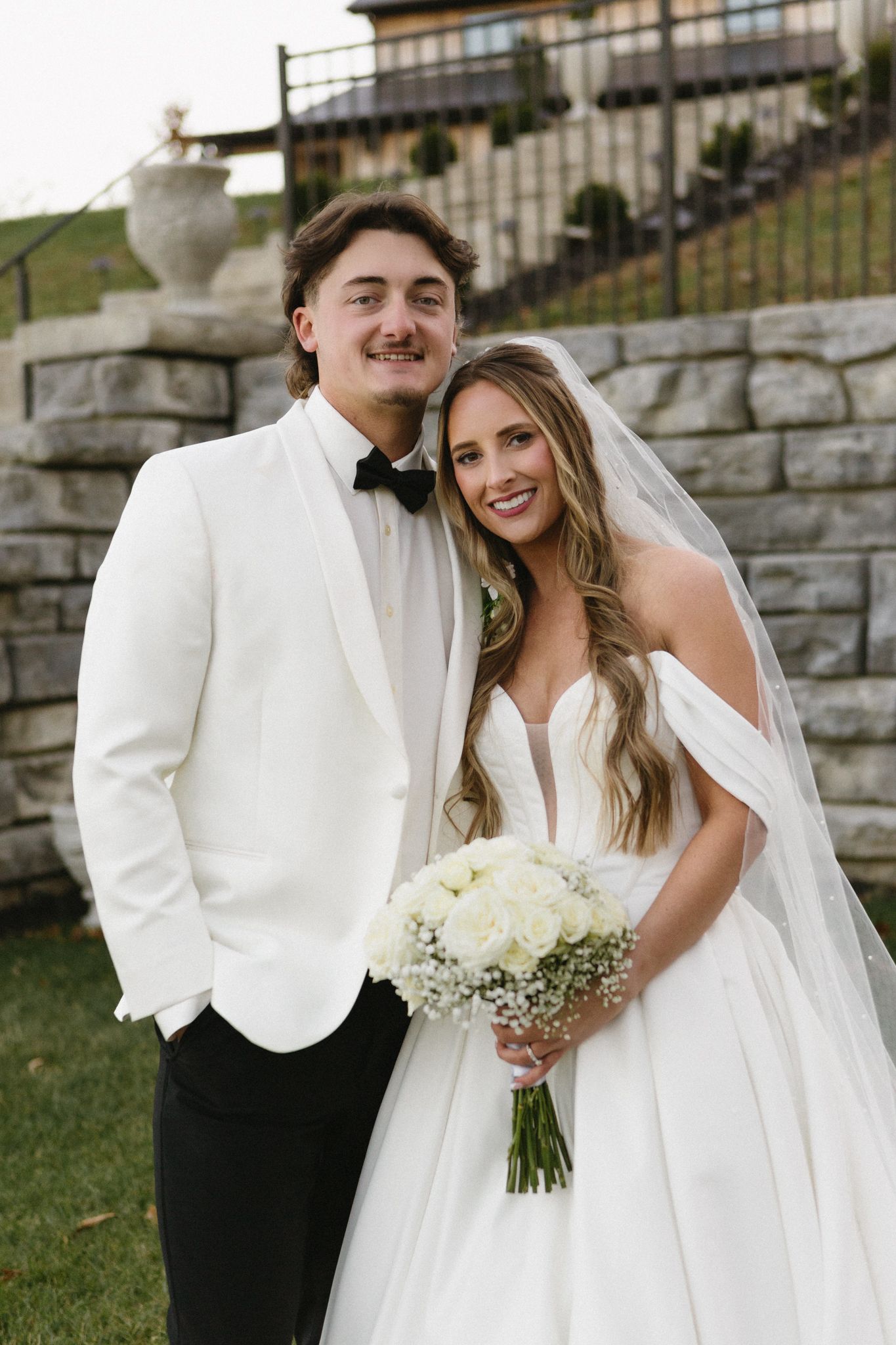 A newlywed couple poses outdoors by stone walls, holding a white floral bouquet.