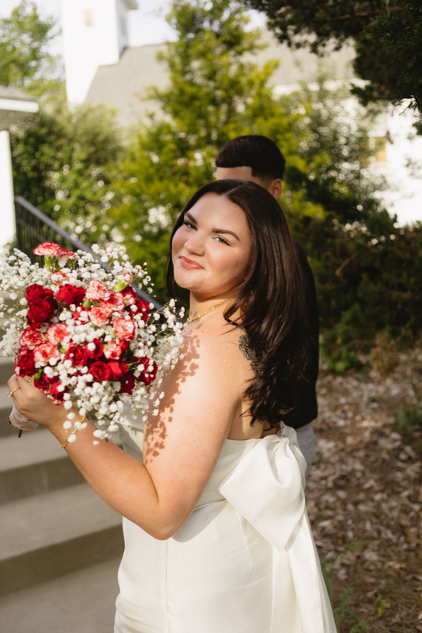 A bride in a white dress holds a beautiful red and white floral bouquet in a romantic outdoor setting.
