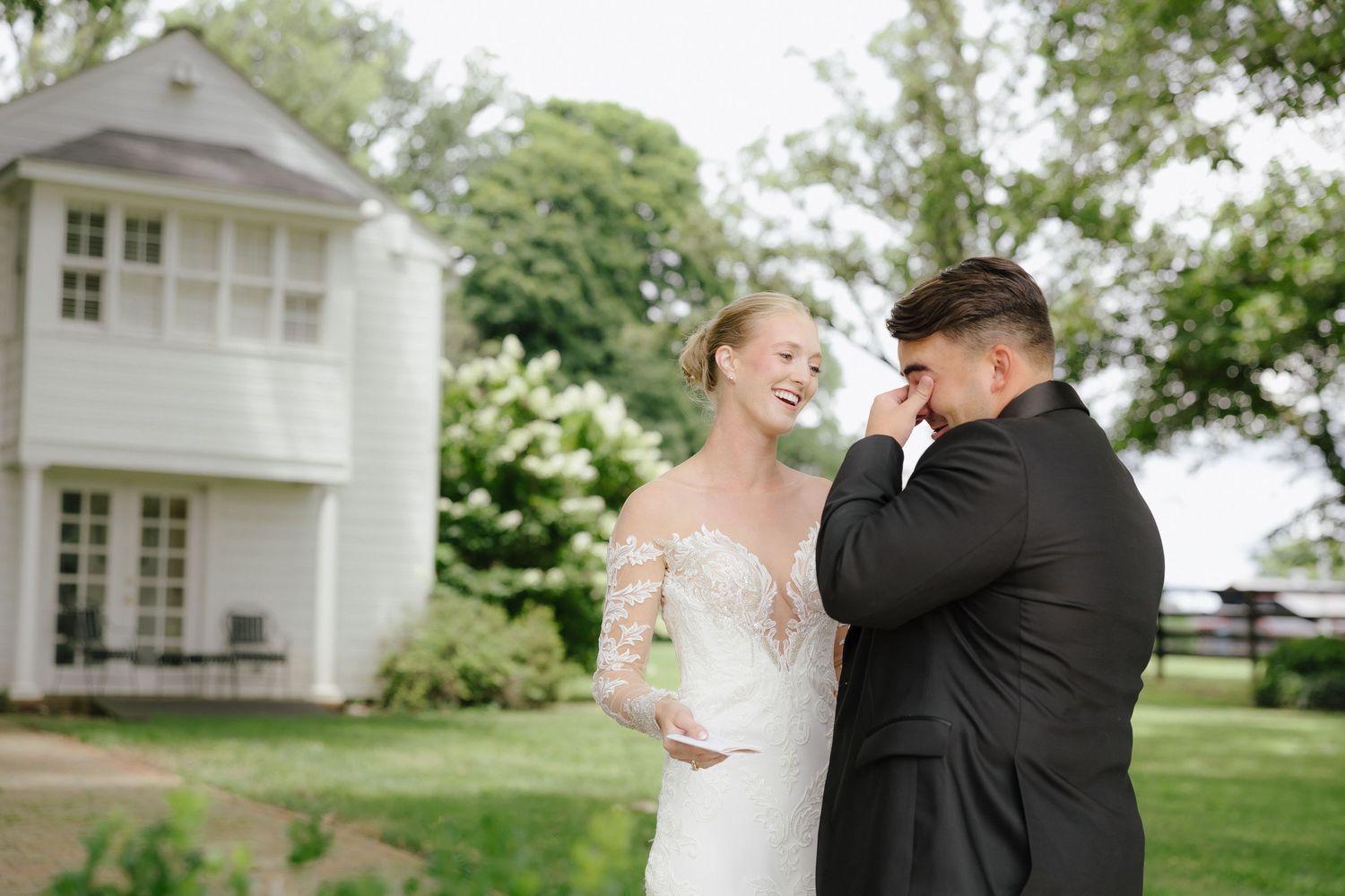 Emotional groom wiping tears during first look with smiling bride in white lace gown outside white farmhouse.