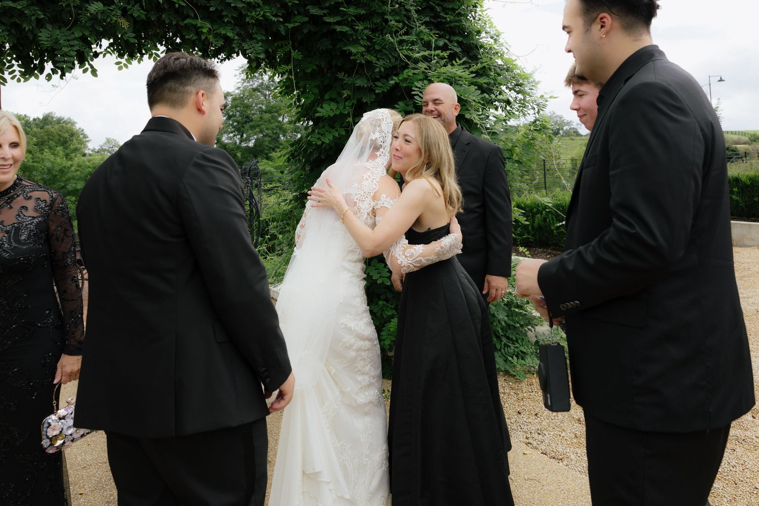 Bride in lace gown and veil embraces guest in black dress at outdoor wedding celebration.