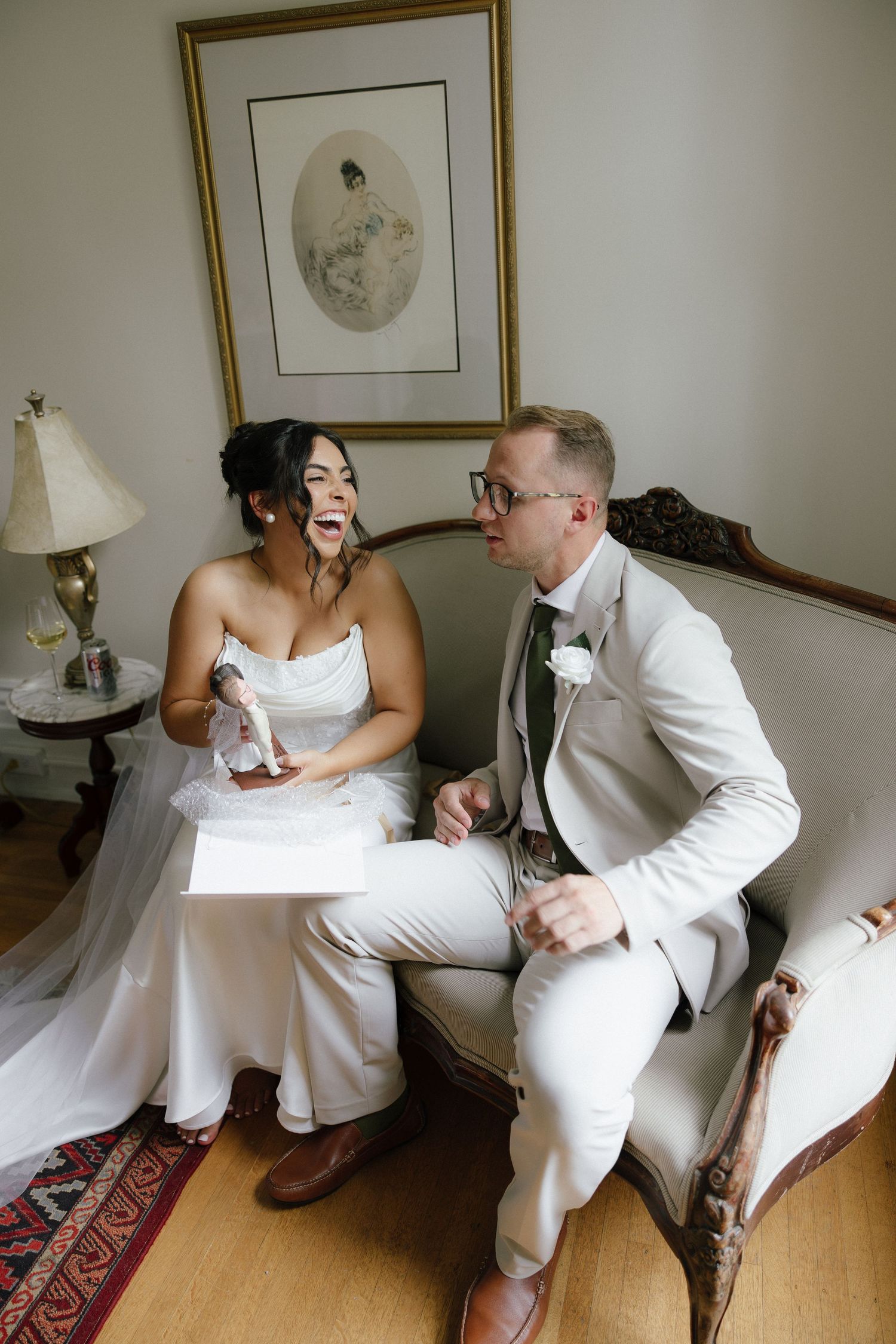Bride and groom laugh together on a vintage sofa, sharing a gift and letter in an elegantly decorated room.