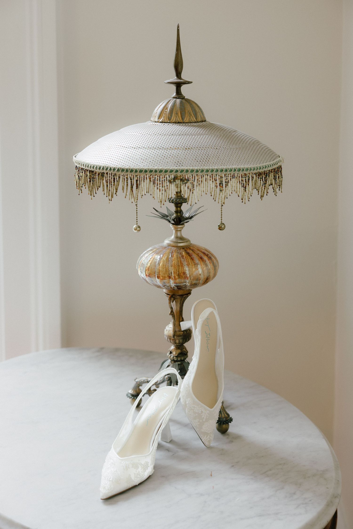 Vintage ornate brass table lamp with beaded fringe shade beside white lace bridal heels on marble table.