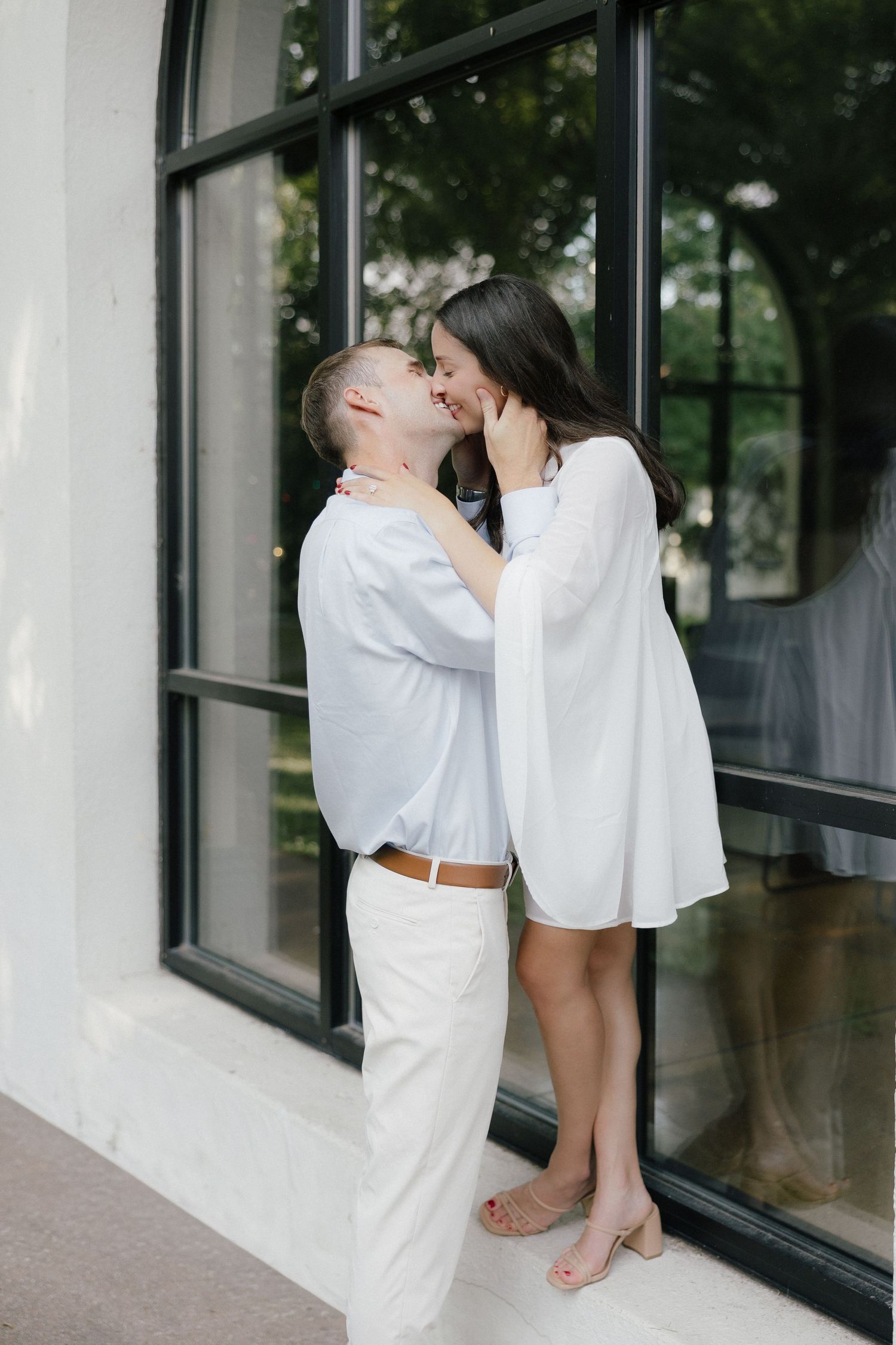 Couple sharing a tender kiss in front of large arched windows, woman in white dress on tiptoes.