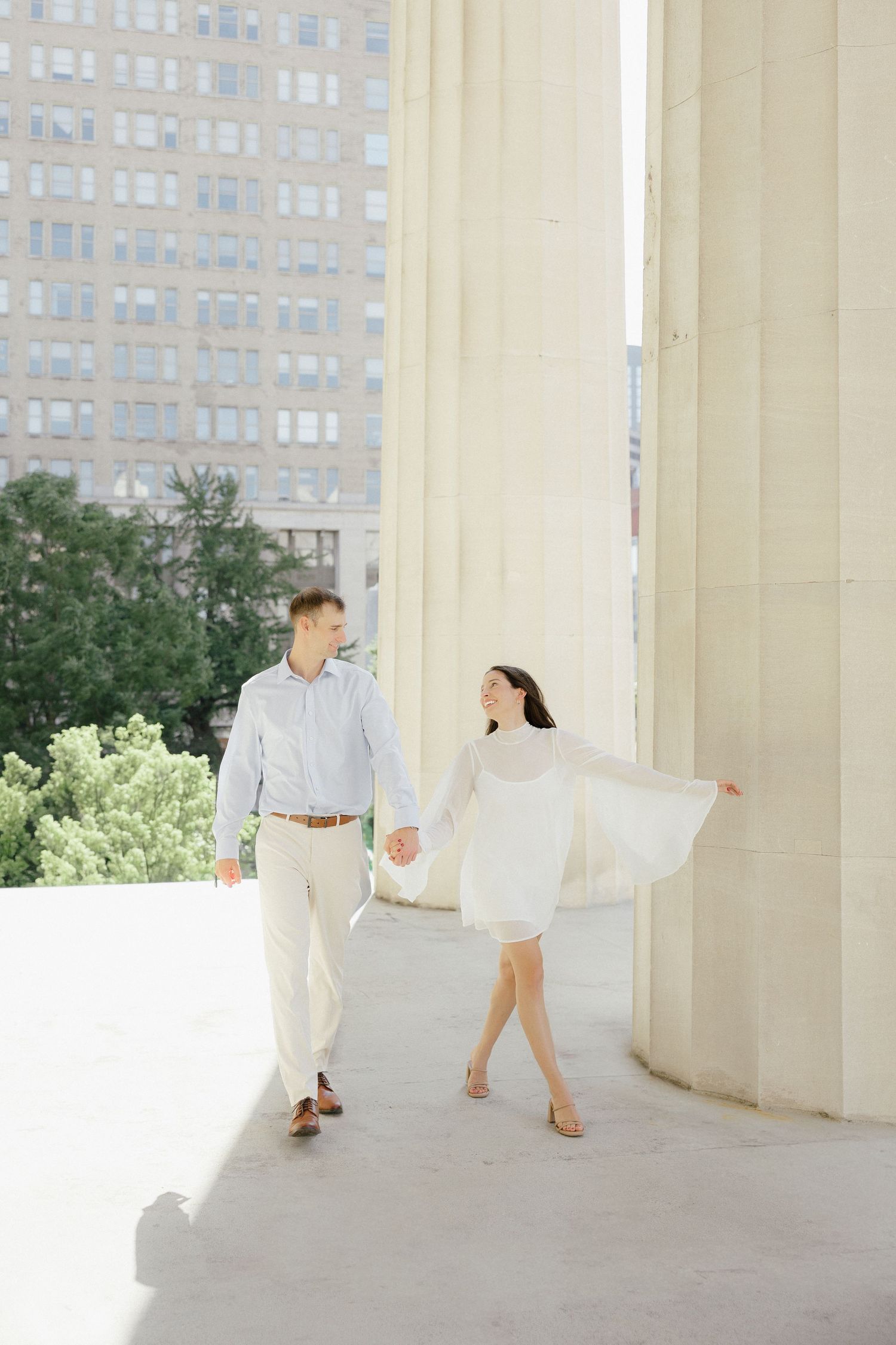 Couple holding hands walking between large white columns with city buildings visible in background.
