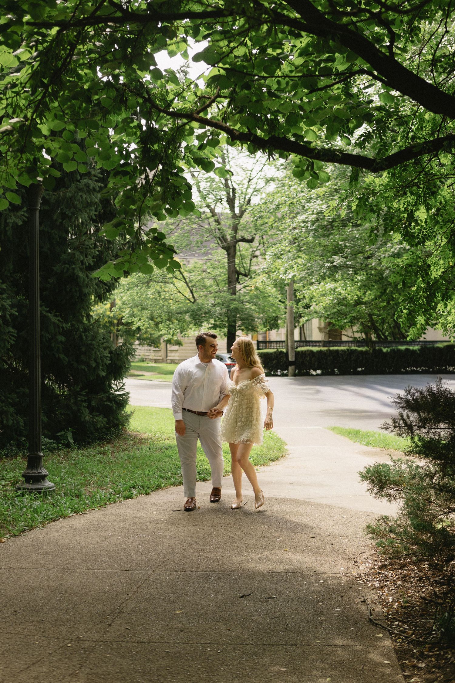 Couple holding hands and walking along a shaded park pathway surrounded by lush green trees.