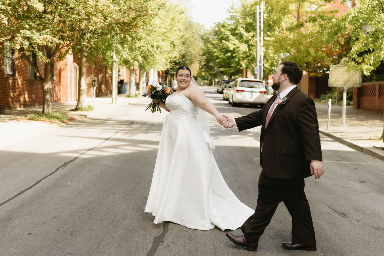 A wedding couple strolls along a tree-lined street during an autumn day.