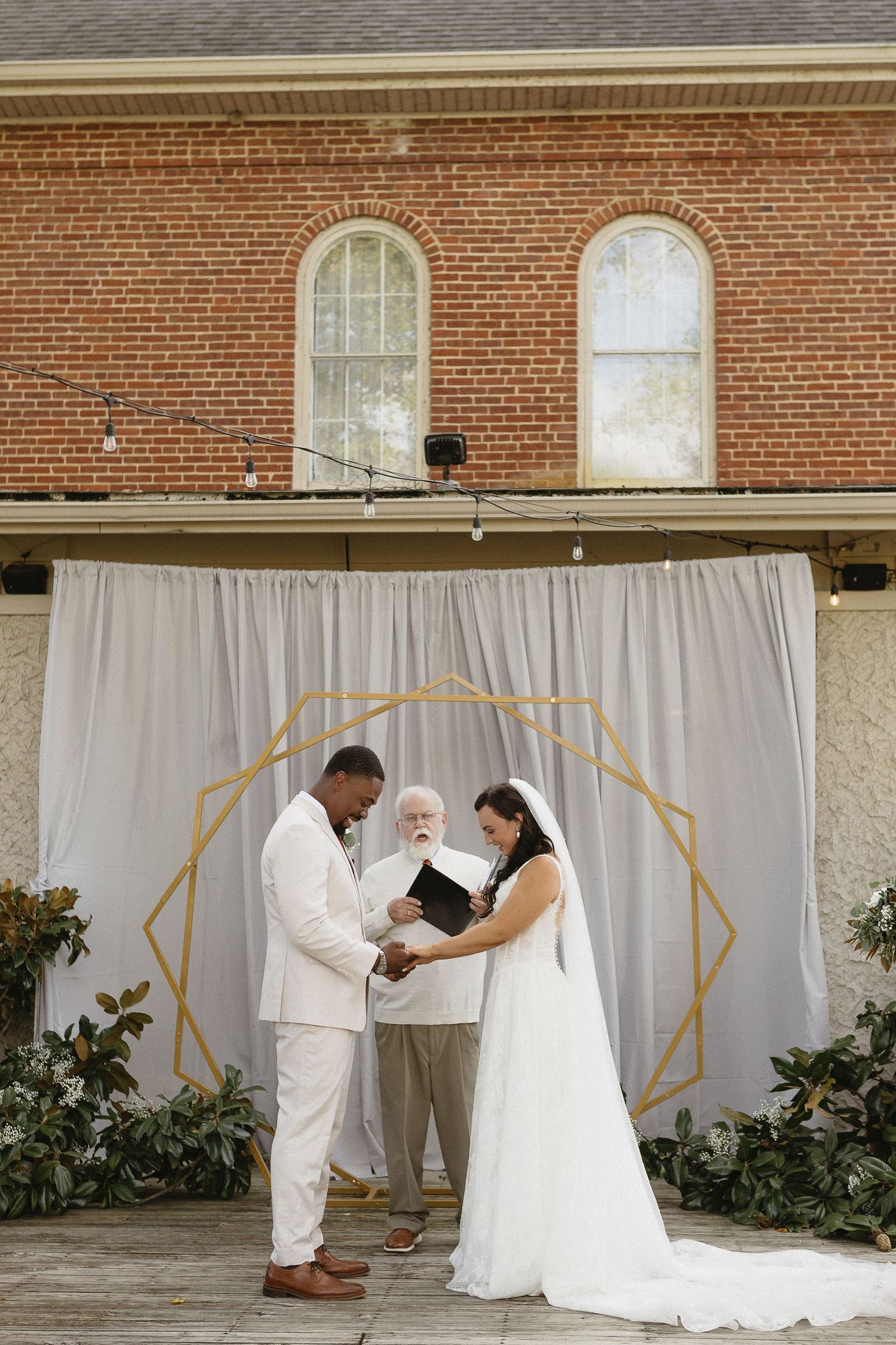 Wedding ceremony in progress with white draping and gold geometric arch backdrop.