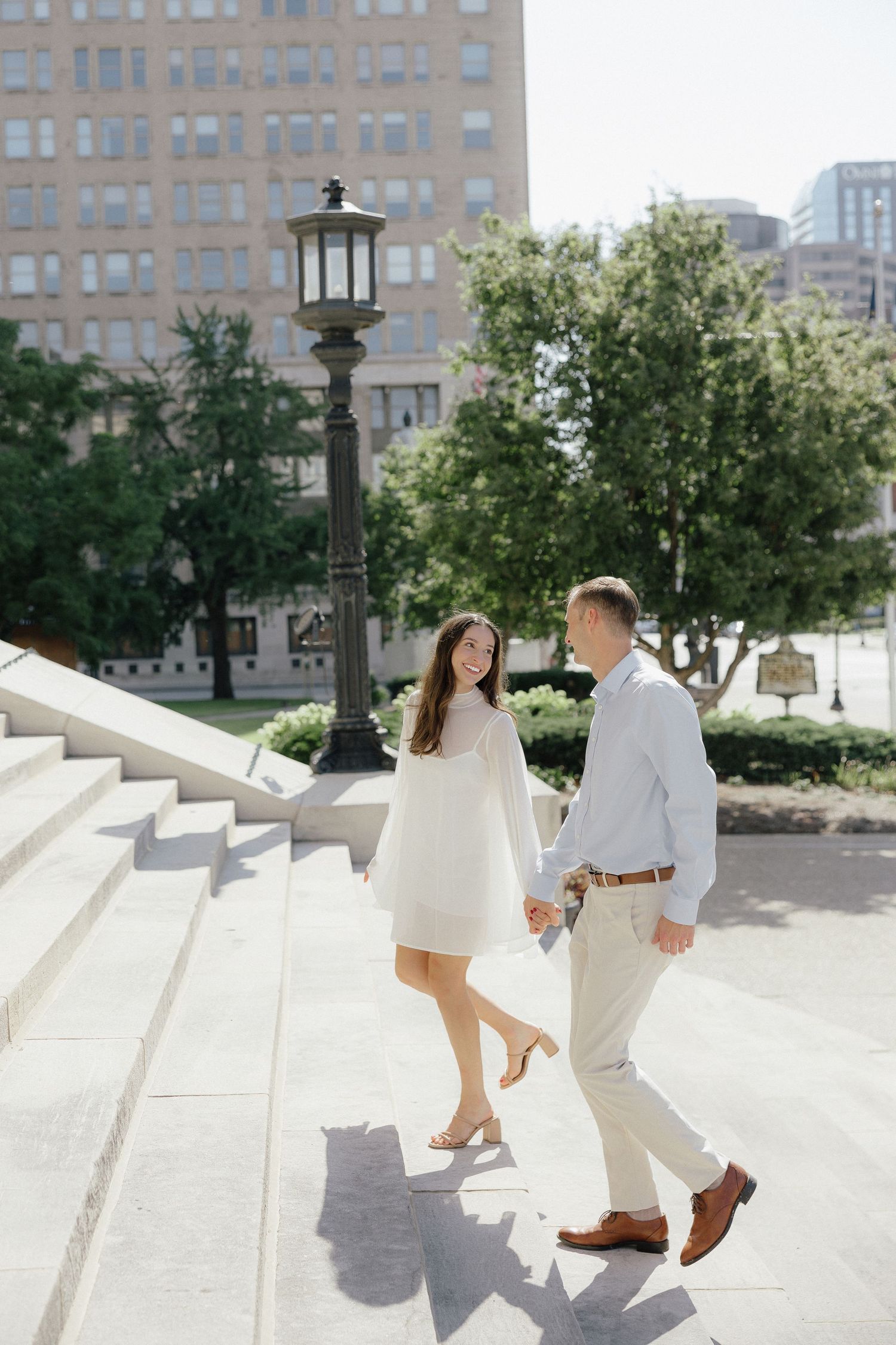 Engaged couple holding hands while walking up stone steps in an urban park setting on a sunny day.