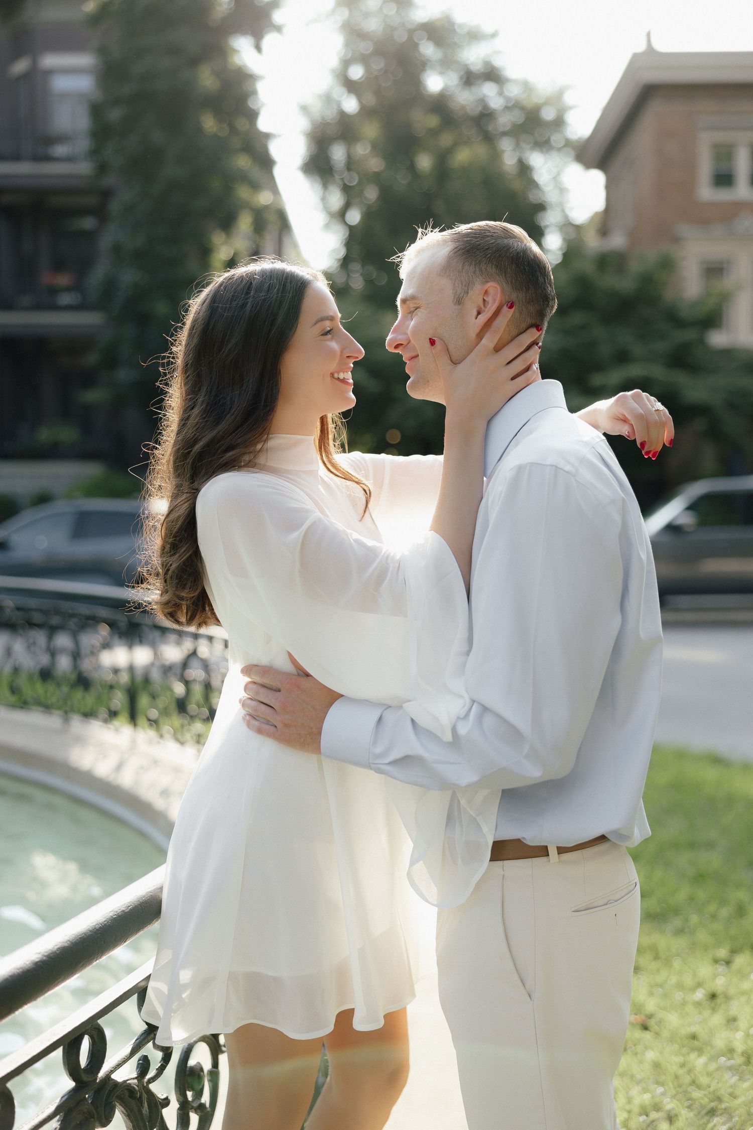 Smiling couple embracing near a fountain in a sunny park, woman in white dress touching mans face.