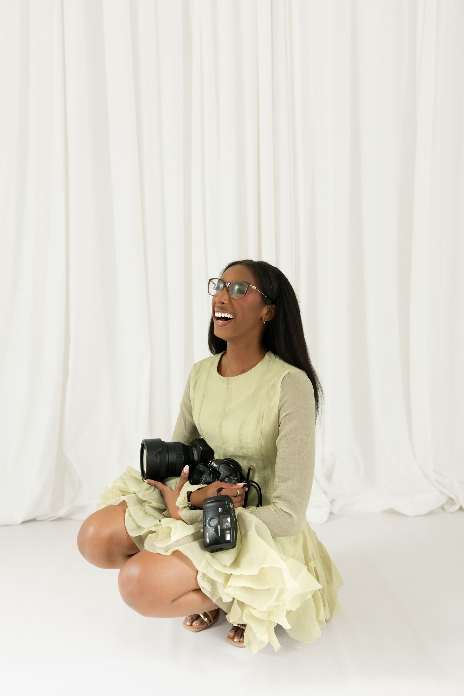 A smiling Black woman photographer sits cross-legged on a white studio floor, holding DSLR cameras, wearing a sage green dress and glasses.