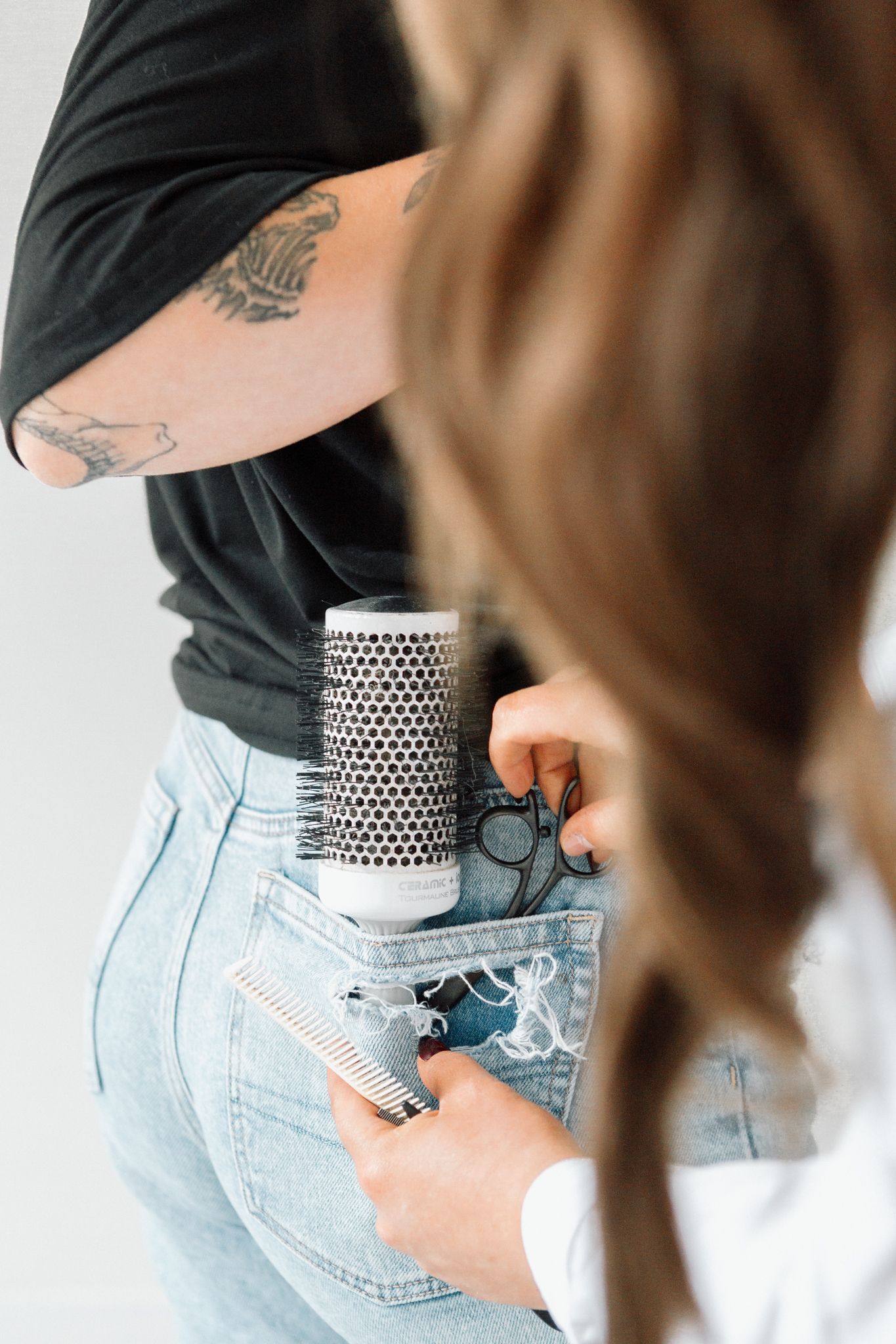 Hairstylist with tattoos storing brushes, scissors, and combs in denim jeans pockets at a hair salon.
