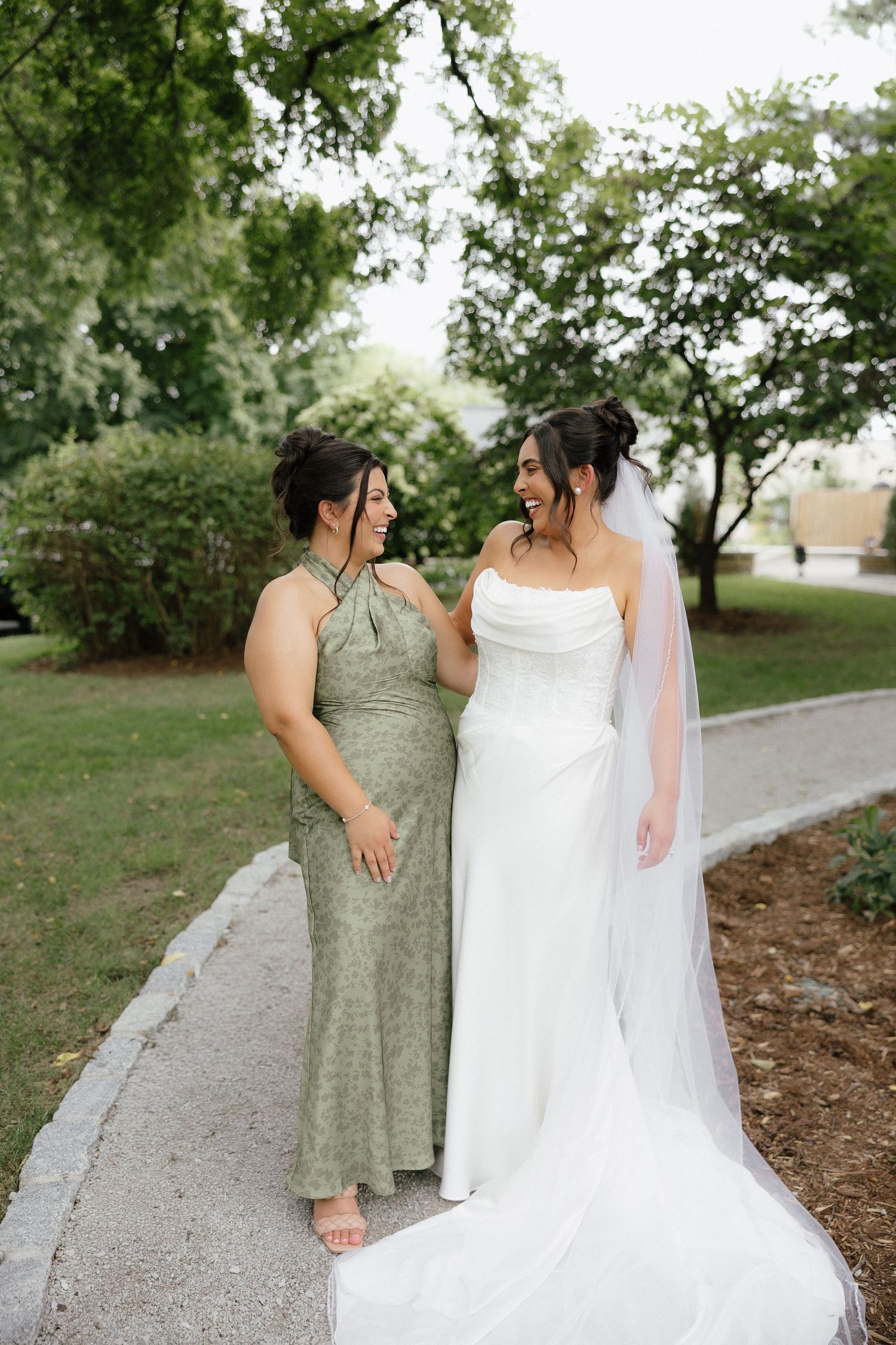 Laughing bride in white gown and bridesmaid in sage green halter dress walking together outdoors.