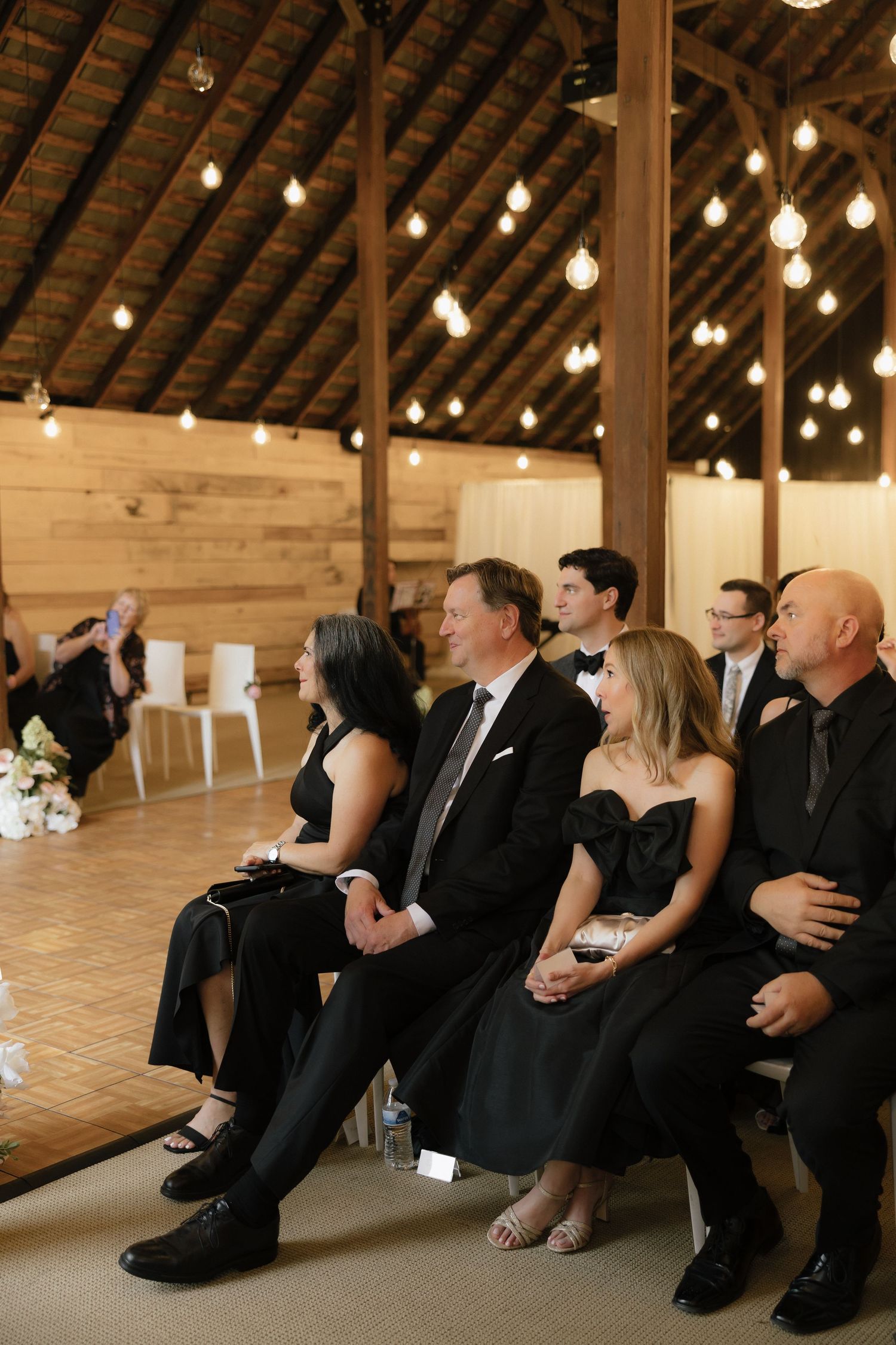 Wedding guests dressed in black attire seated during barn venue ceremony with warm Edison bulb lights.