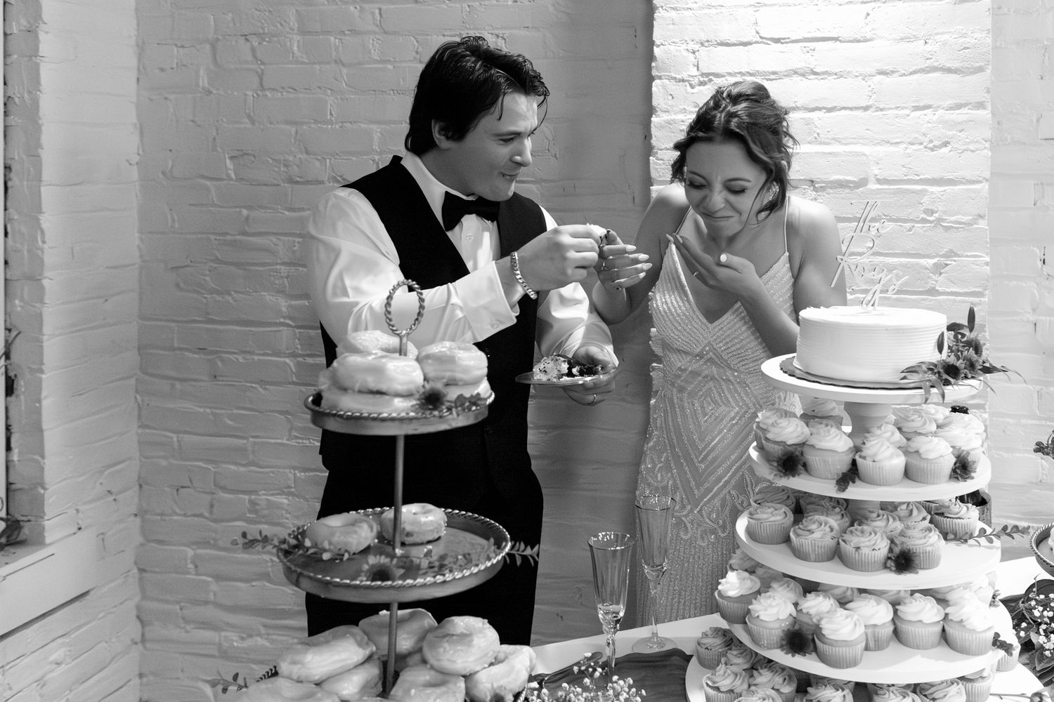 A black and white photo of a wedding couple enjoying their dessert display with cupcakes and cake tiers at their reception.