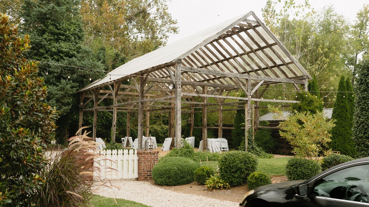 A wooden pergola structure with ornate beams stands in a landscaped garden with trimmed bushes and trees. Garden Fall Wedding venue.