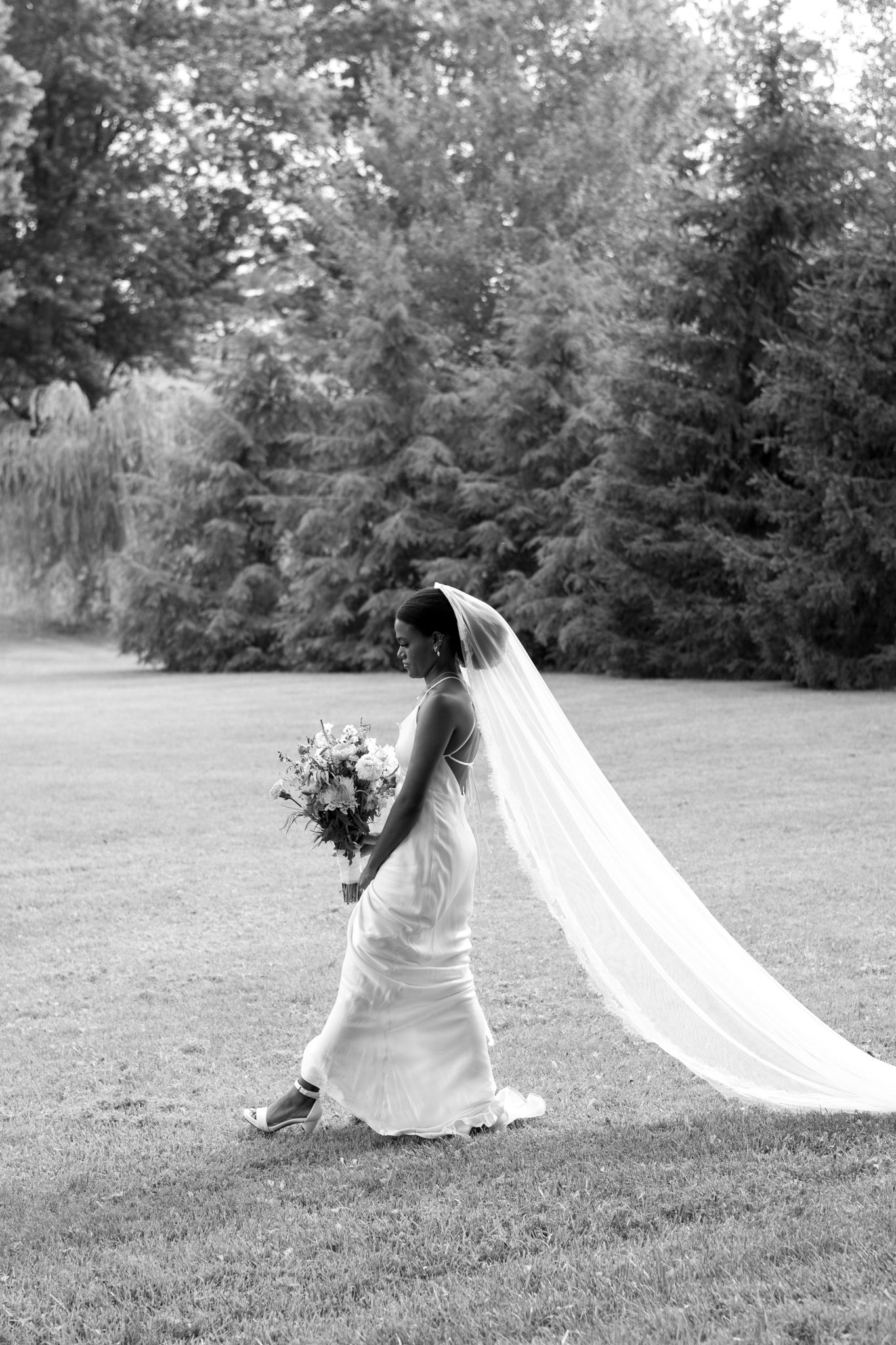 A flowing white wedding veil billows dramatically across a grassy field in black and white photography.