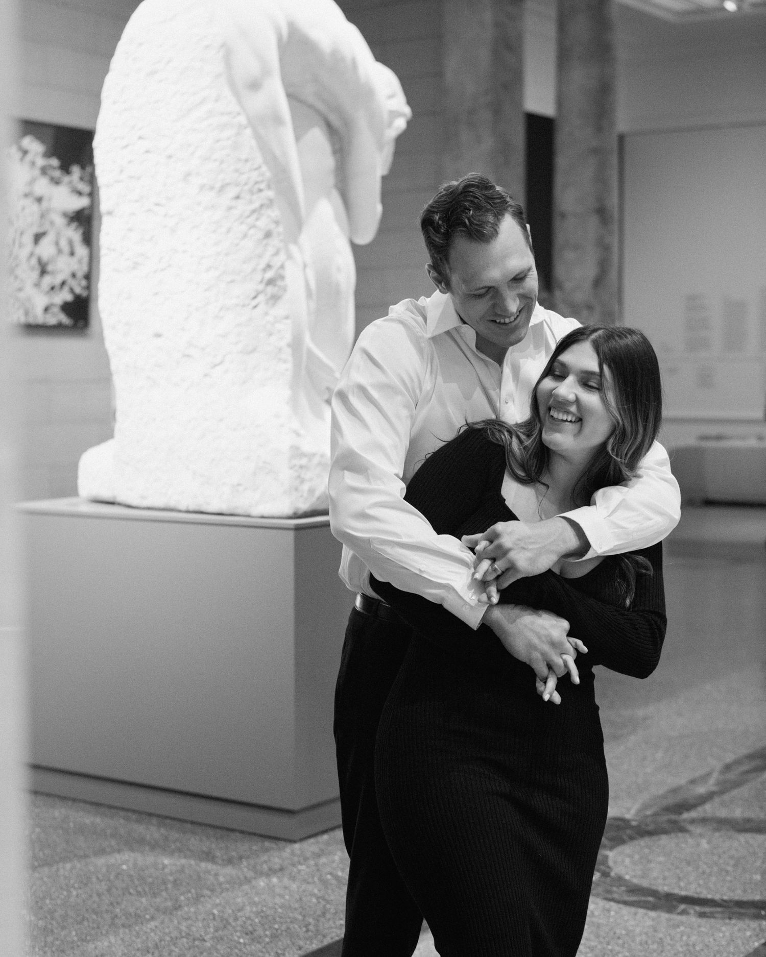 Engaged couple in downtown Louisville, with the woman smiling and showing her engagement ring while embracing her fiancé