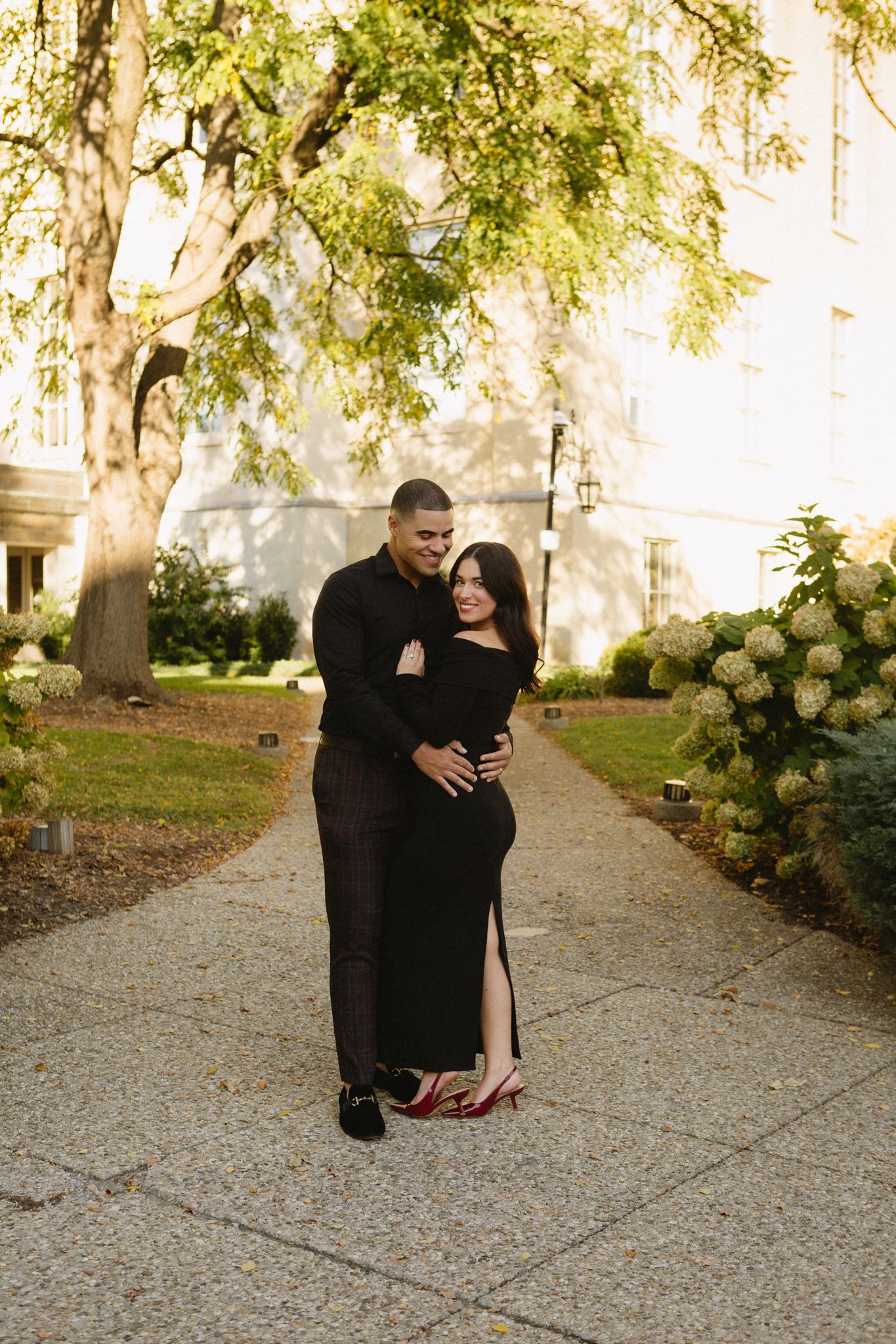 A couple in matching black formal attire embrace on a garden path lined with trees and hydrangeas at sunset.