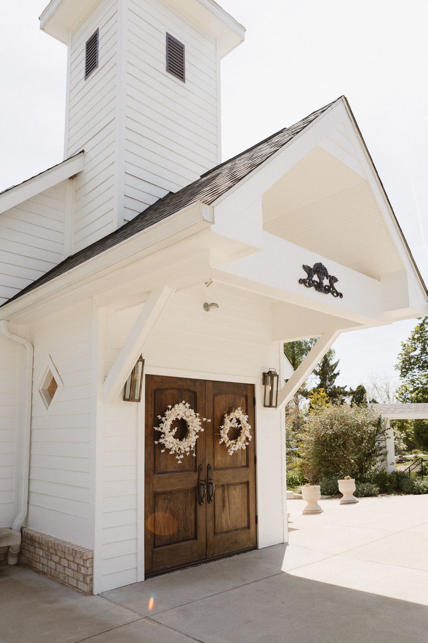 A charming white chapel entrance with wooden double doors adorned with matching wreaths and featuring a tall bell tower.