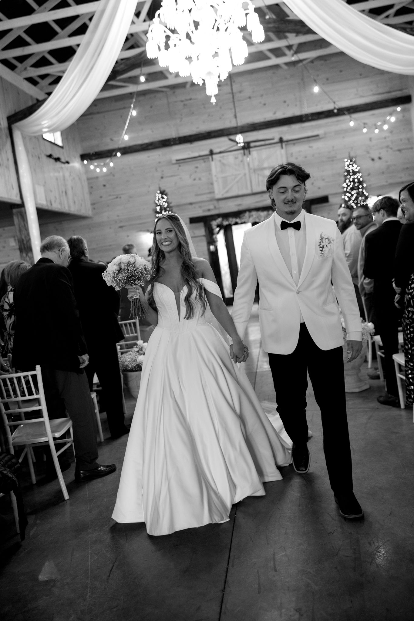 A black and white photo of newlyweds walking down the aisle under crystal chandeliers and white draping.