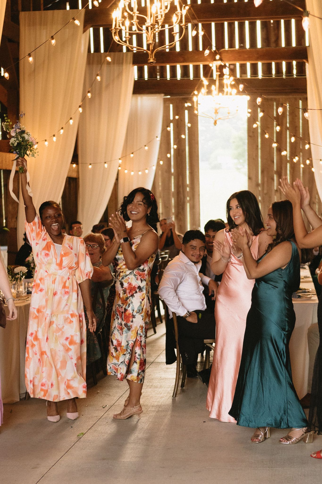 Wedding guests dance and celebrate in a barn venue decorated with string lights and white drapes.