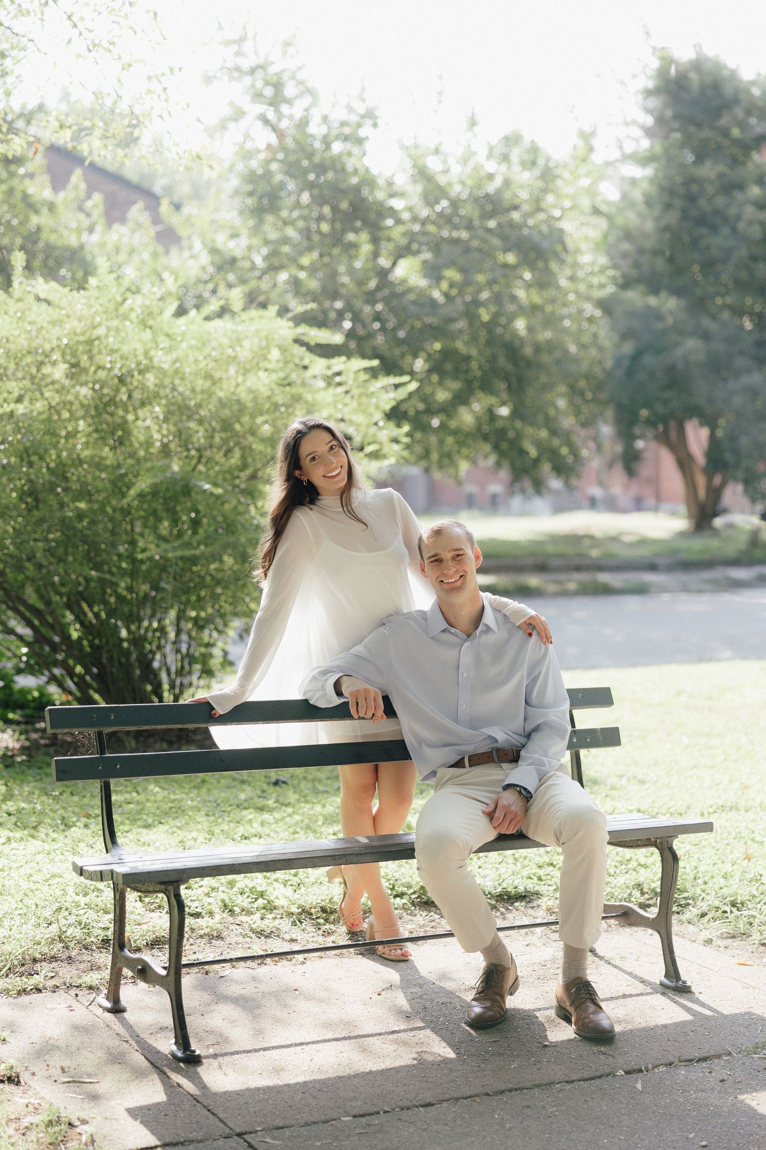 Smiling couple posing in sunny park, woman in white dress standing beside man seated on a bench.