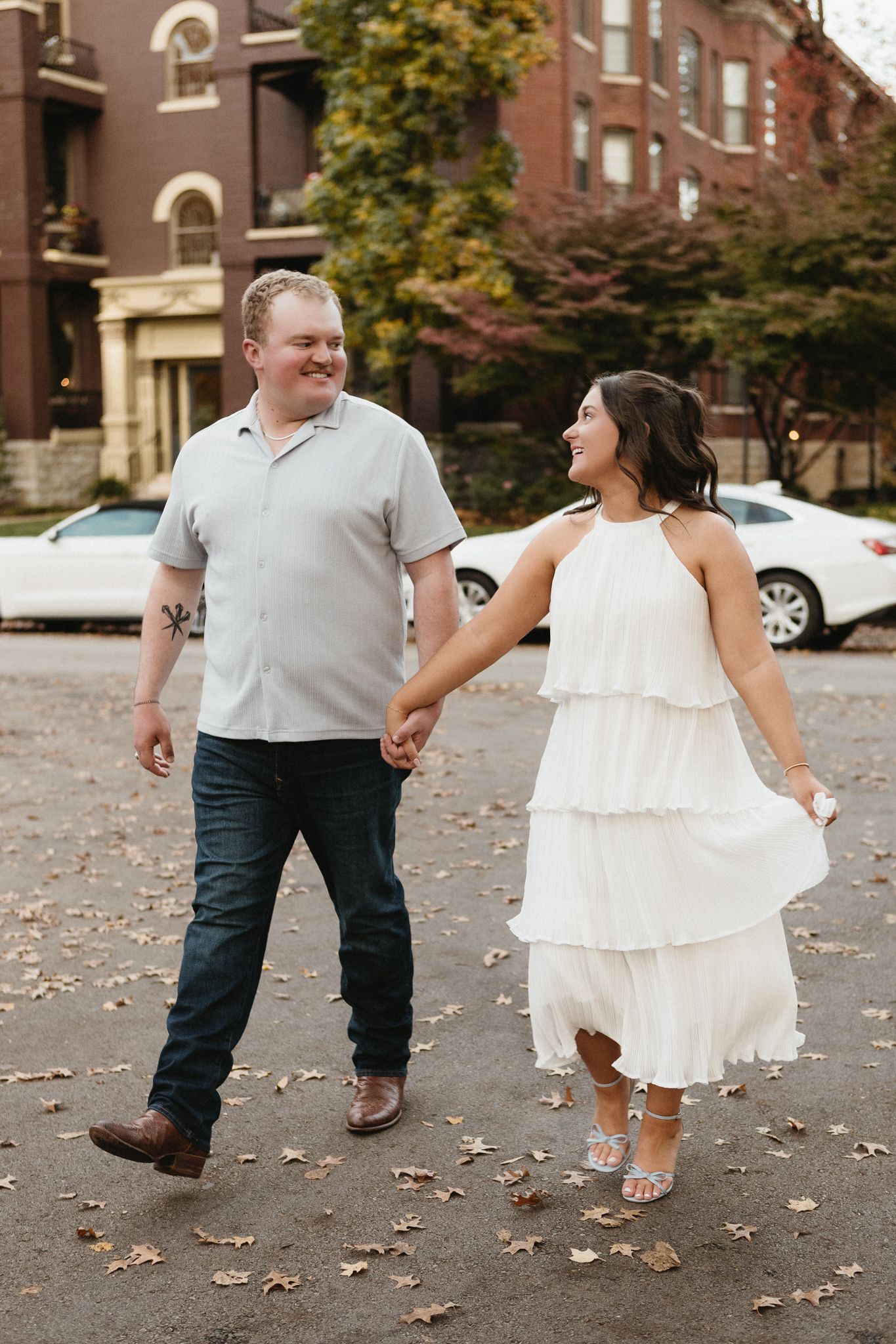 Two people walk together on fallen autumn leaves in front of a brick building.