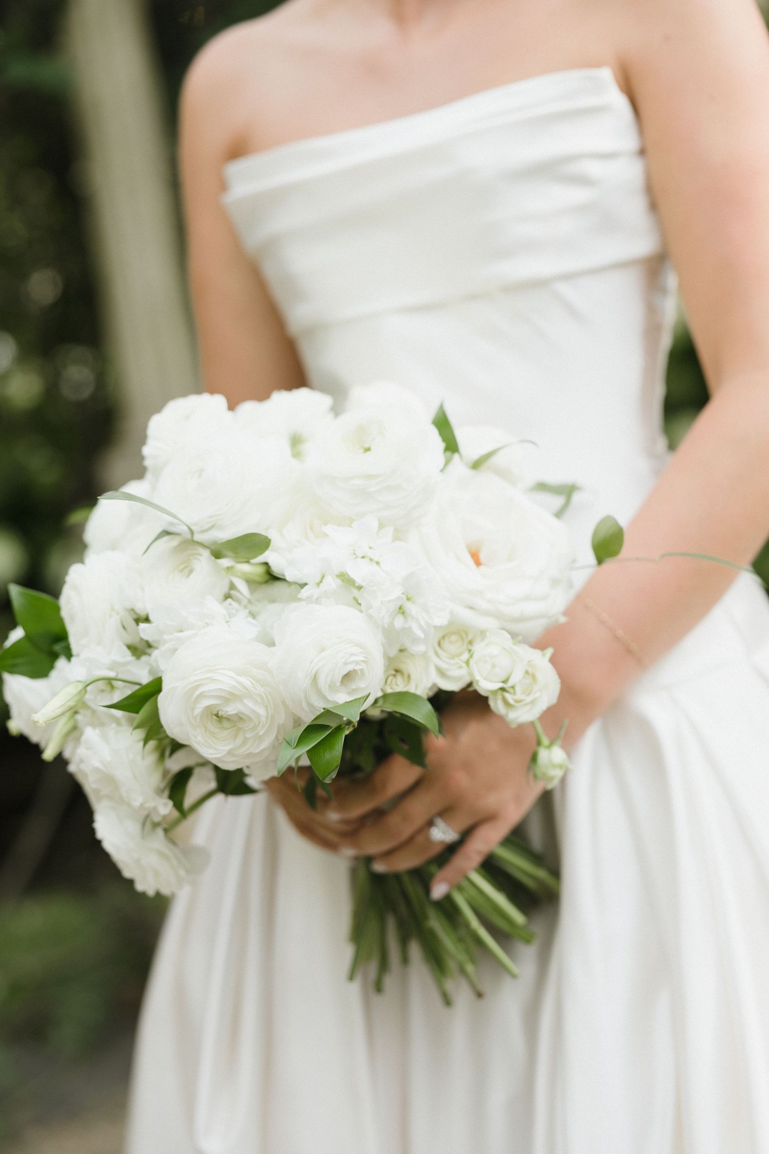 Bride in strapless white gown holding elegant all-white bouquet of ranunculus and roses with green foliage.
